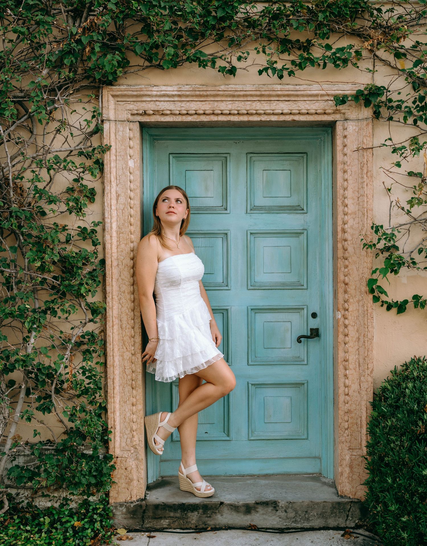 Woman in white strapless mini dress posing by a teal vintage door framed with ivy on a beige wall.