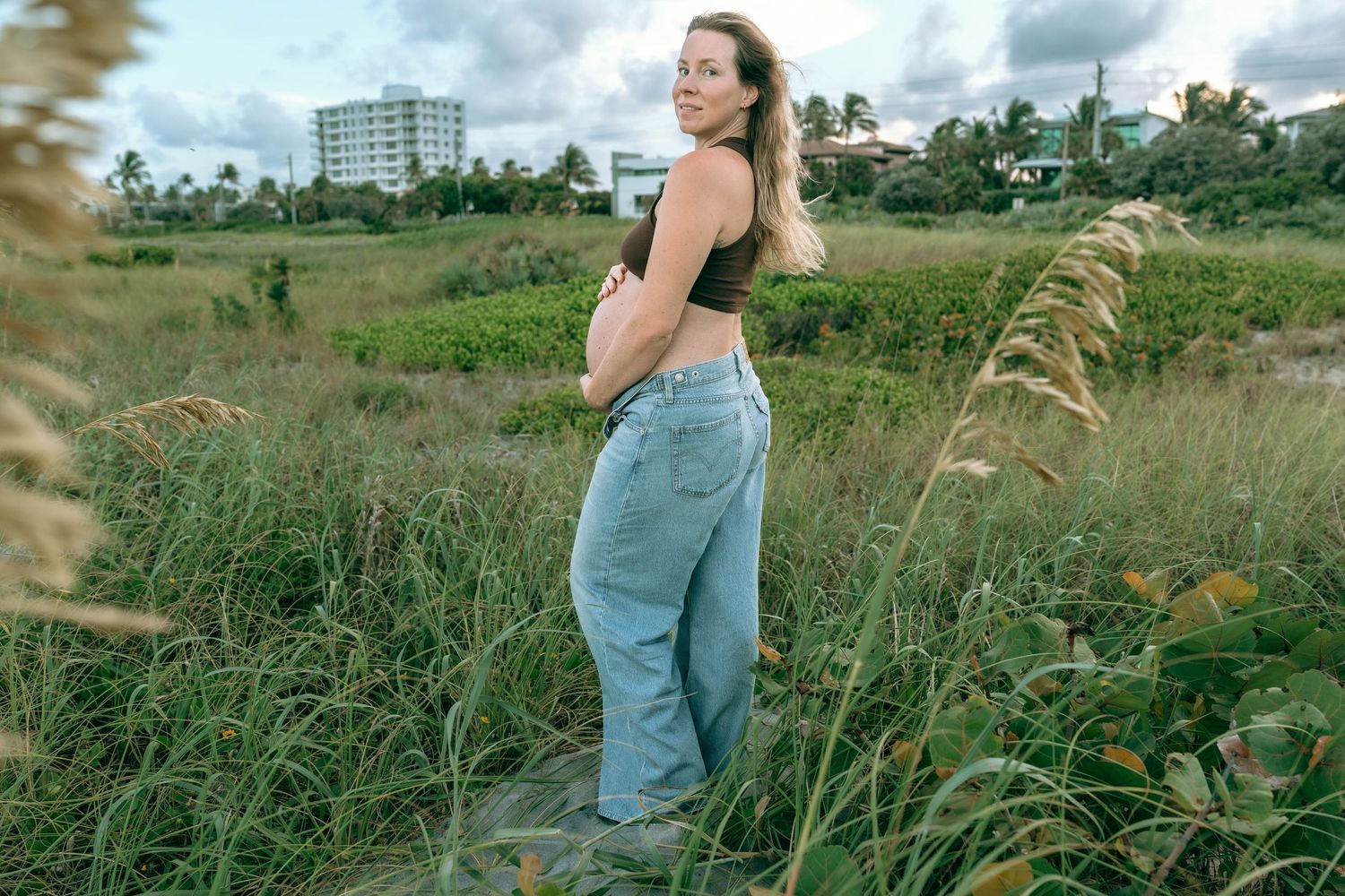 Pregnant woman in crop top and jeans standing in lush green field with tropical buildings in background.
