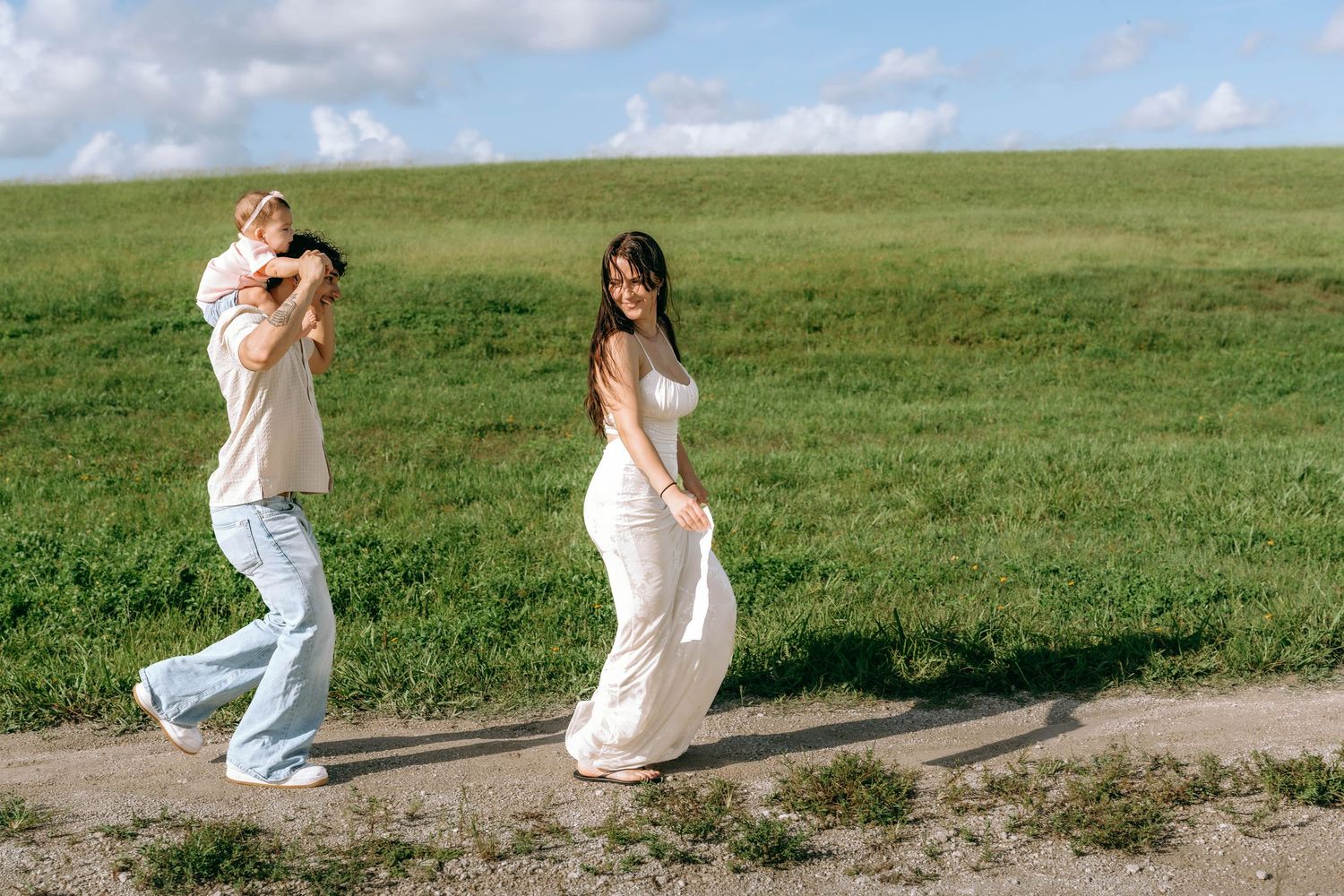Couple walking on a dirt path through green hills, woman in white dress and man in jeans on a sunny day.