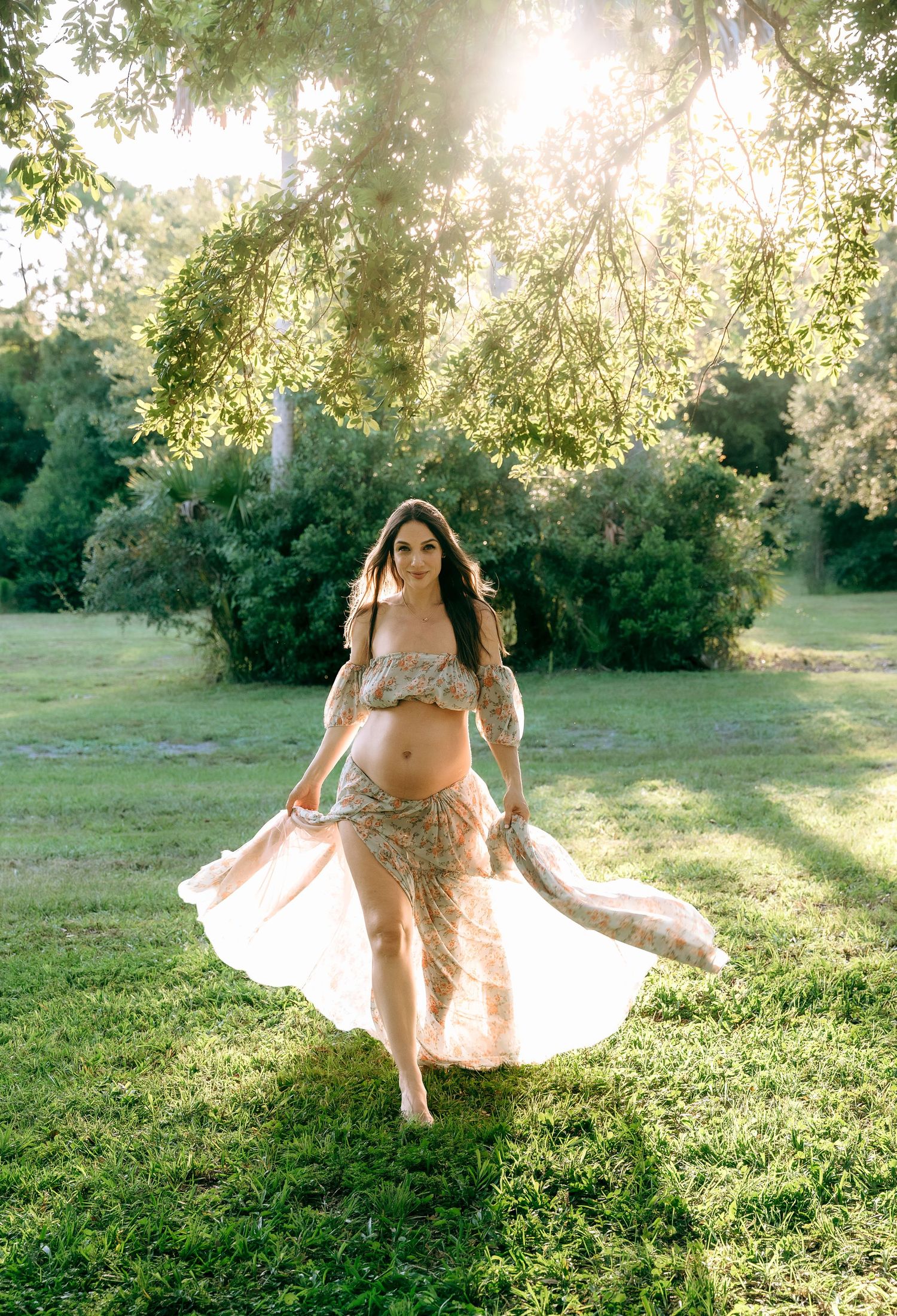 Dancing maternity portraits with flowing dress in sun-dappled meadow.