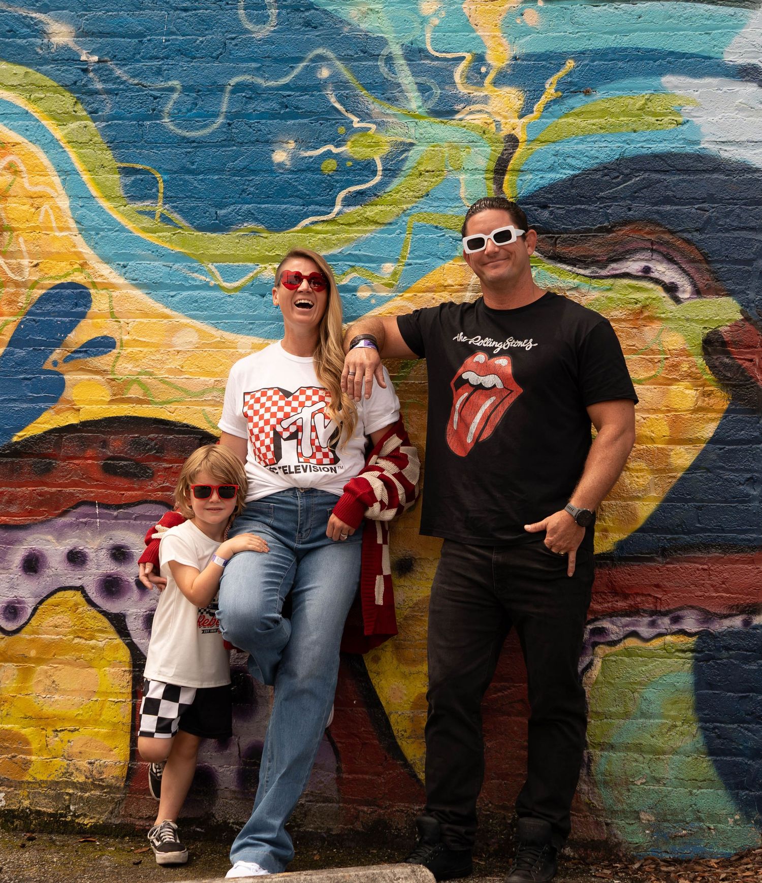 Family of three posing and smiling in front of colorful graffiti mural, wearing Rolling Stones and MTV graphic tees.