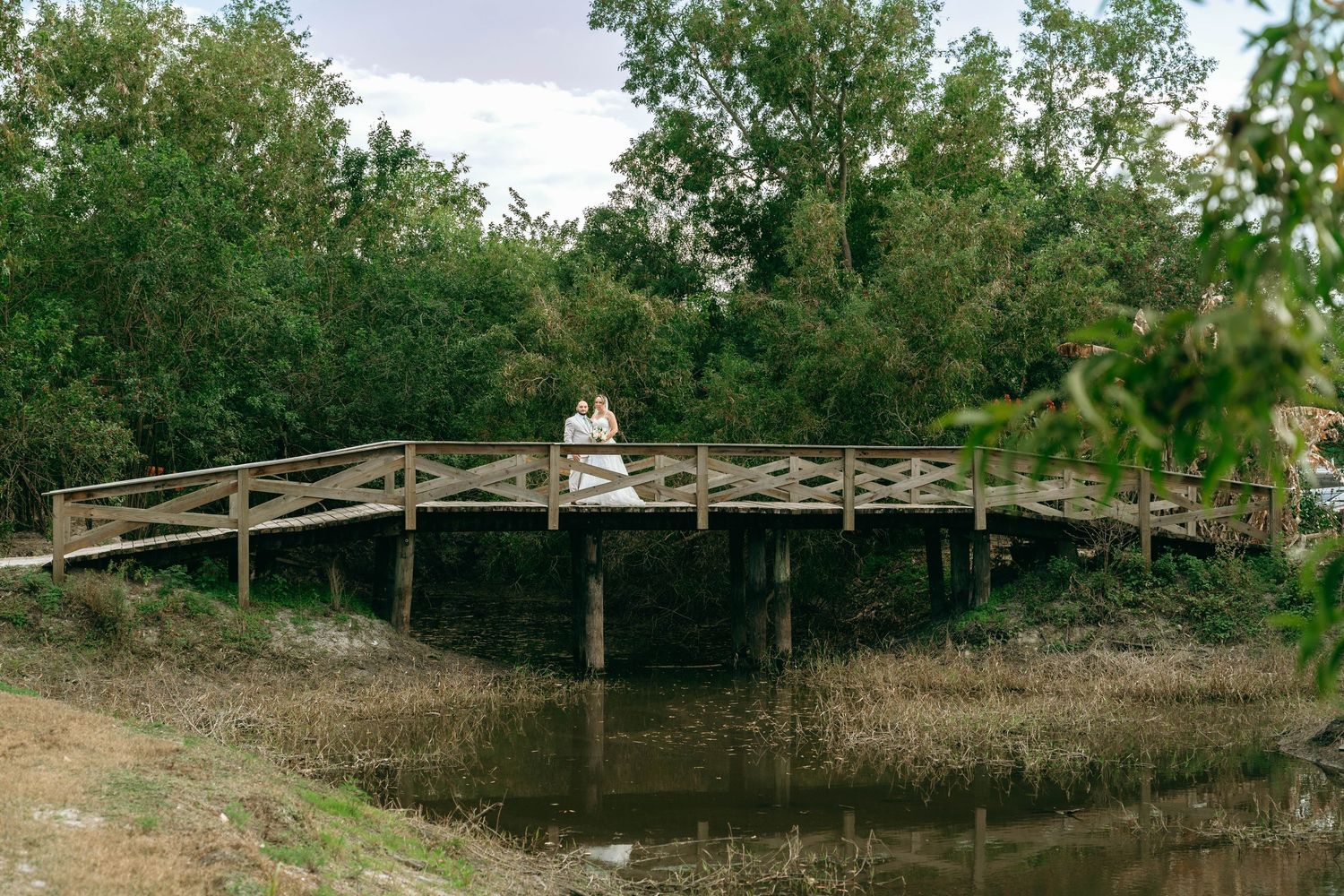 Rustic wooden bridge walkway extends through lush green trees and foliage in a natural outdoor setting.