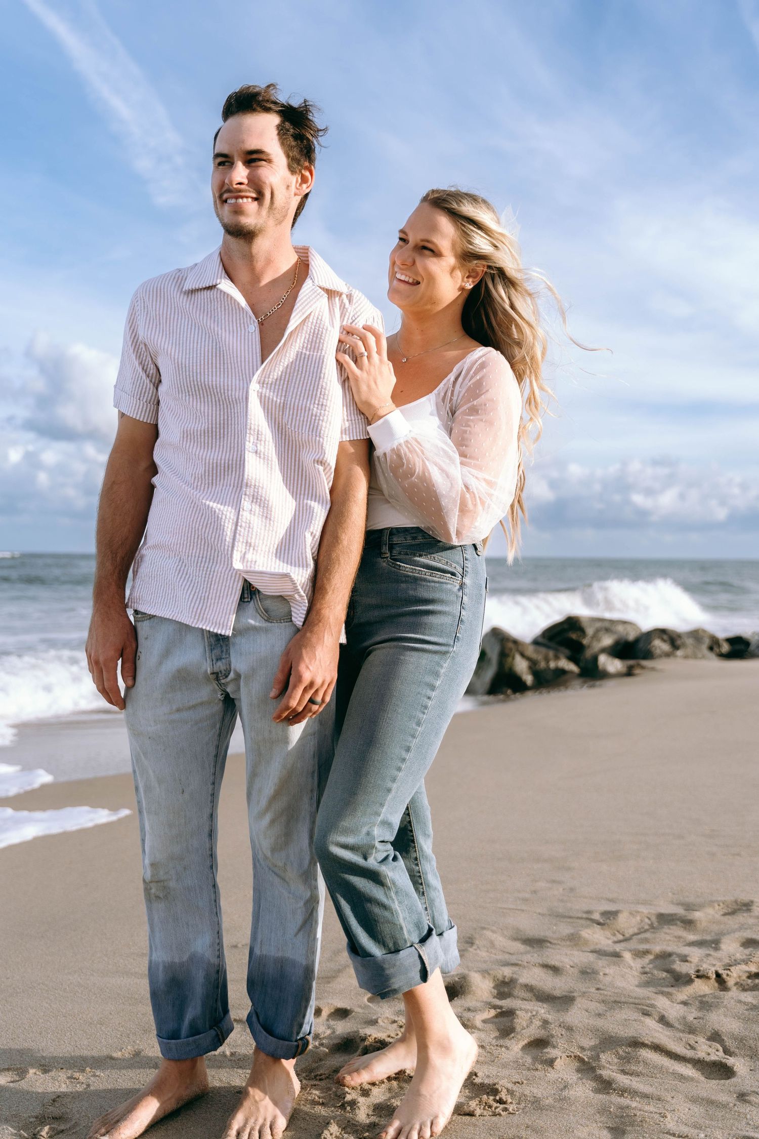 A couple walks playfully together on a sandy beach with ocean waves in the background.