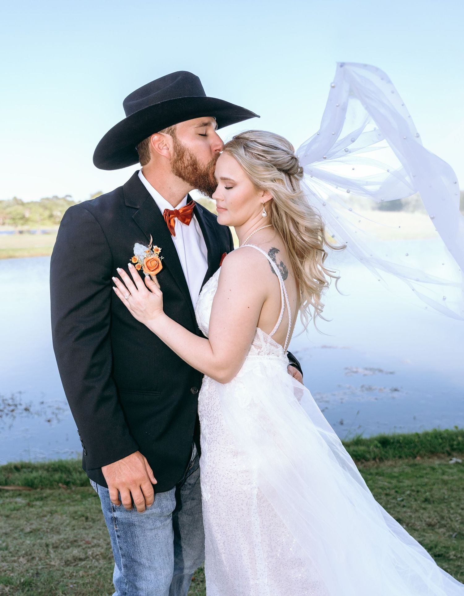A bride and groom share intimate moments as her veil flows in the wind by a lakeside.