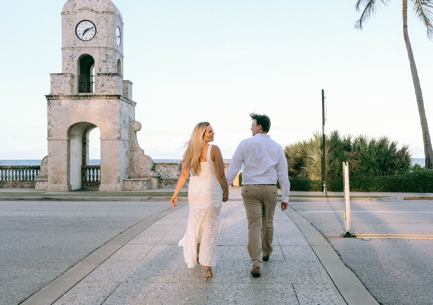 Couple walking hand in hand past a historic clock tower near the ocean.