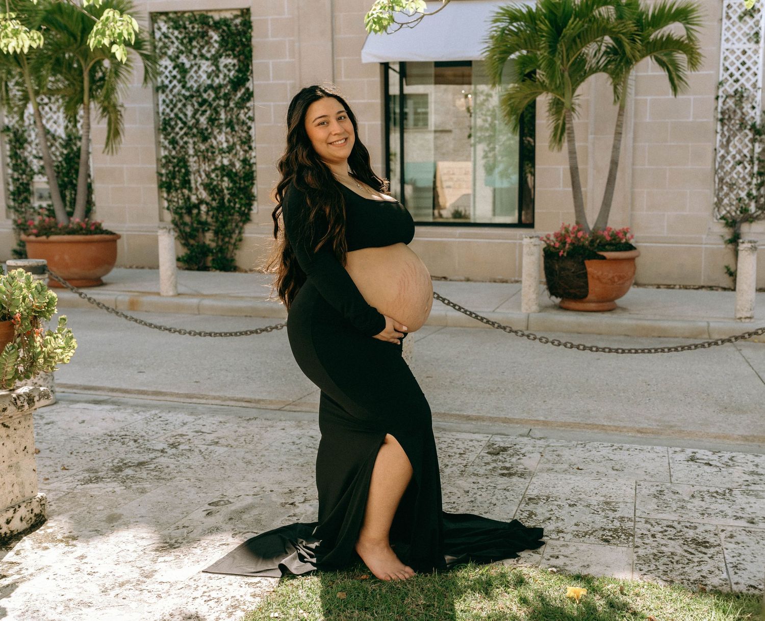 Pregnant woman in elegant black maternity gown poses outdoors cradling baby bump in tropical garden setting. Worth Avenue, Palm Beach, FL