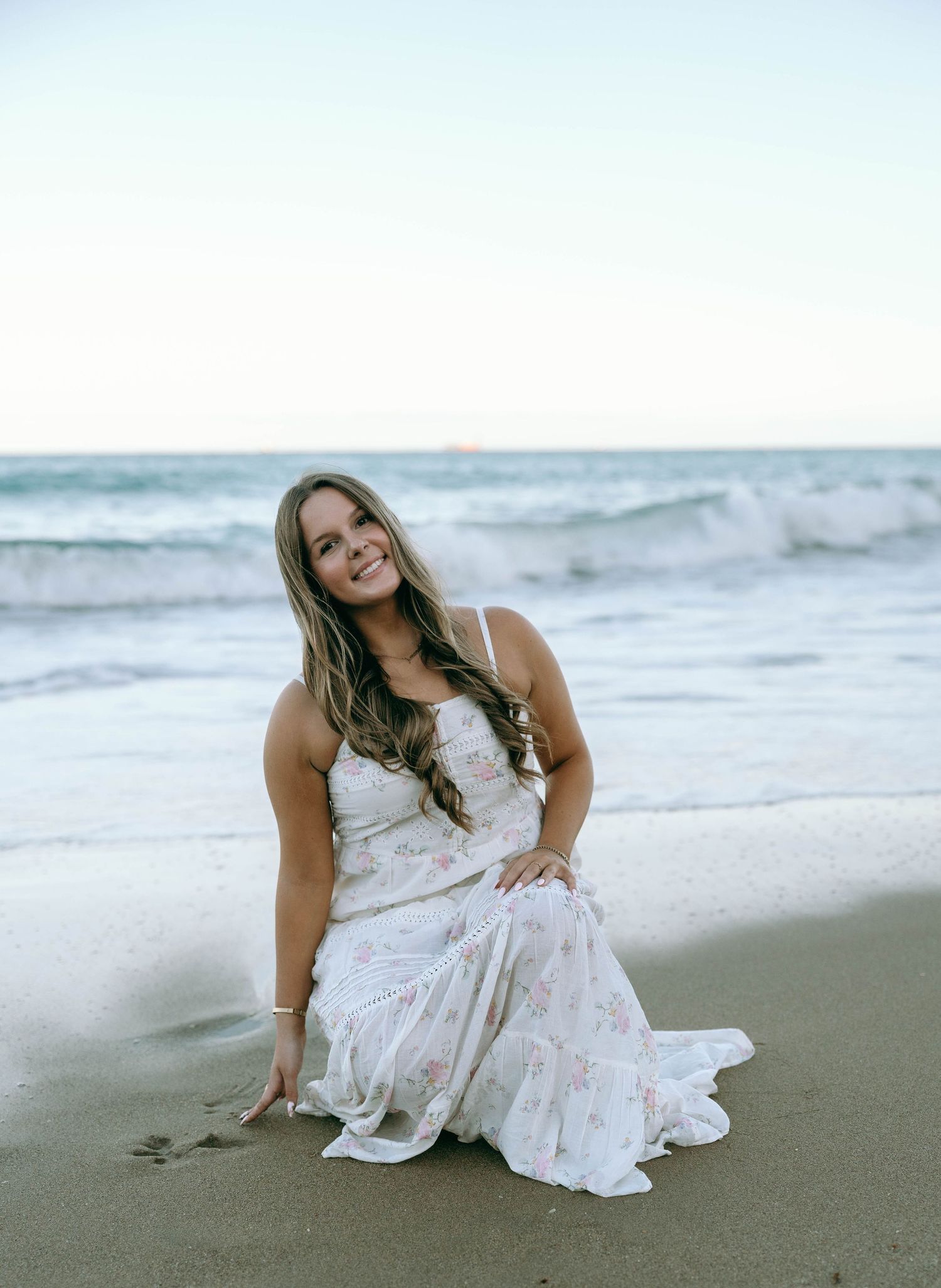 Woman in white floral gown sits on sandy beach with ocean waves at golden hour. Worth Avenue, Palm Beach, FL