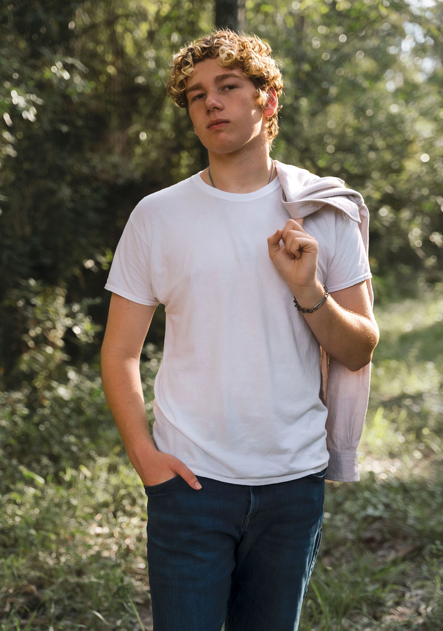 A person in white t-shirt and jeans poses casually in a wooded area.