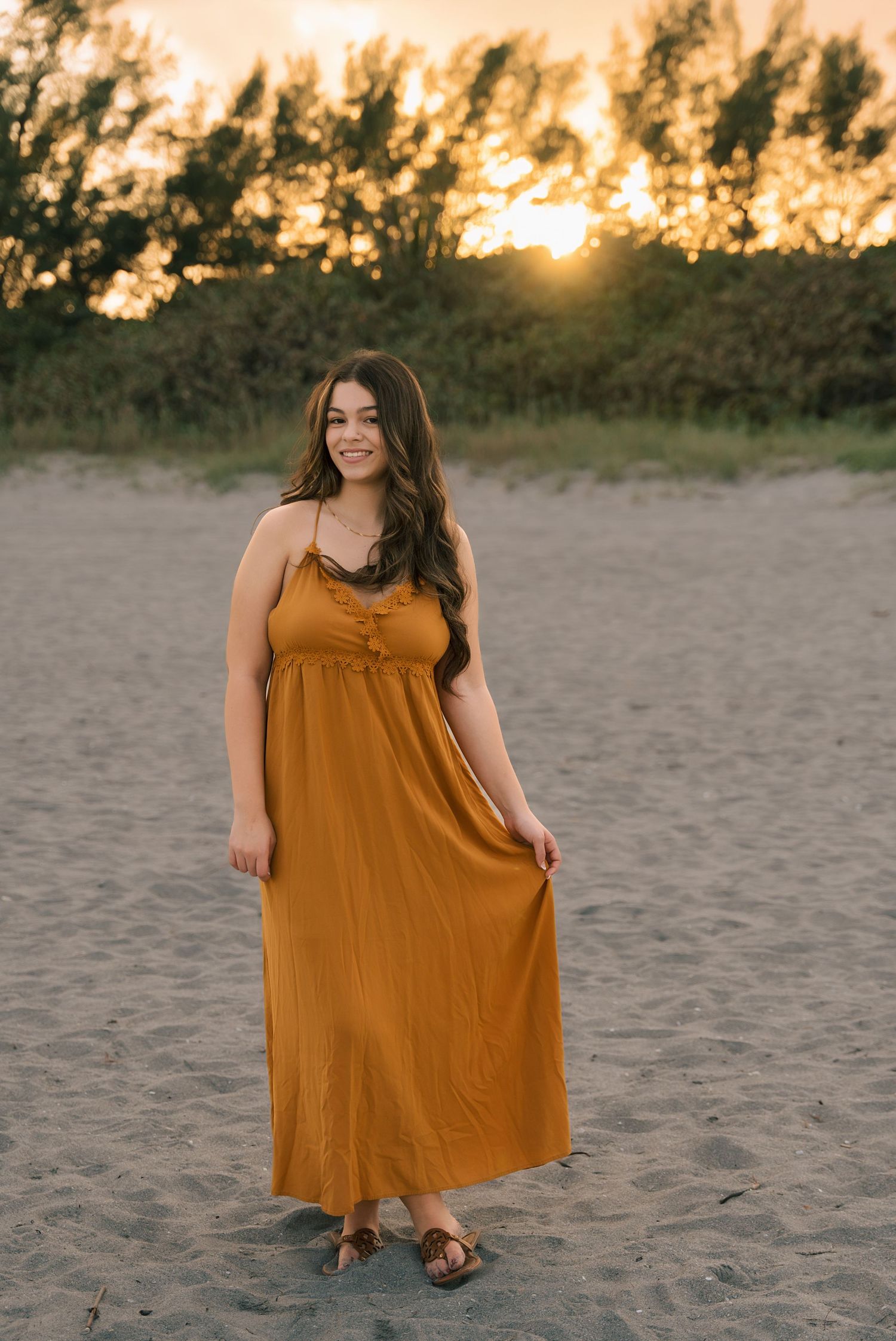 A flowing rust-colored maxi dress photographed at sunset on a sandy beach.