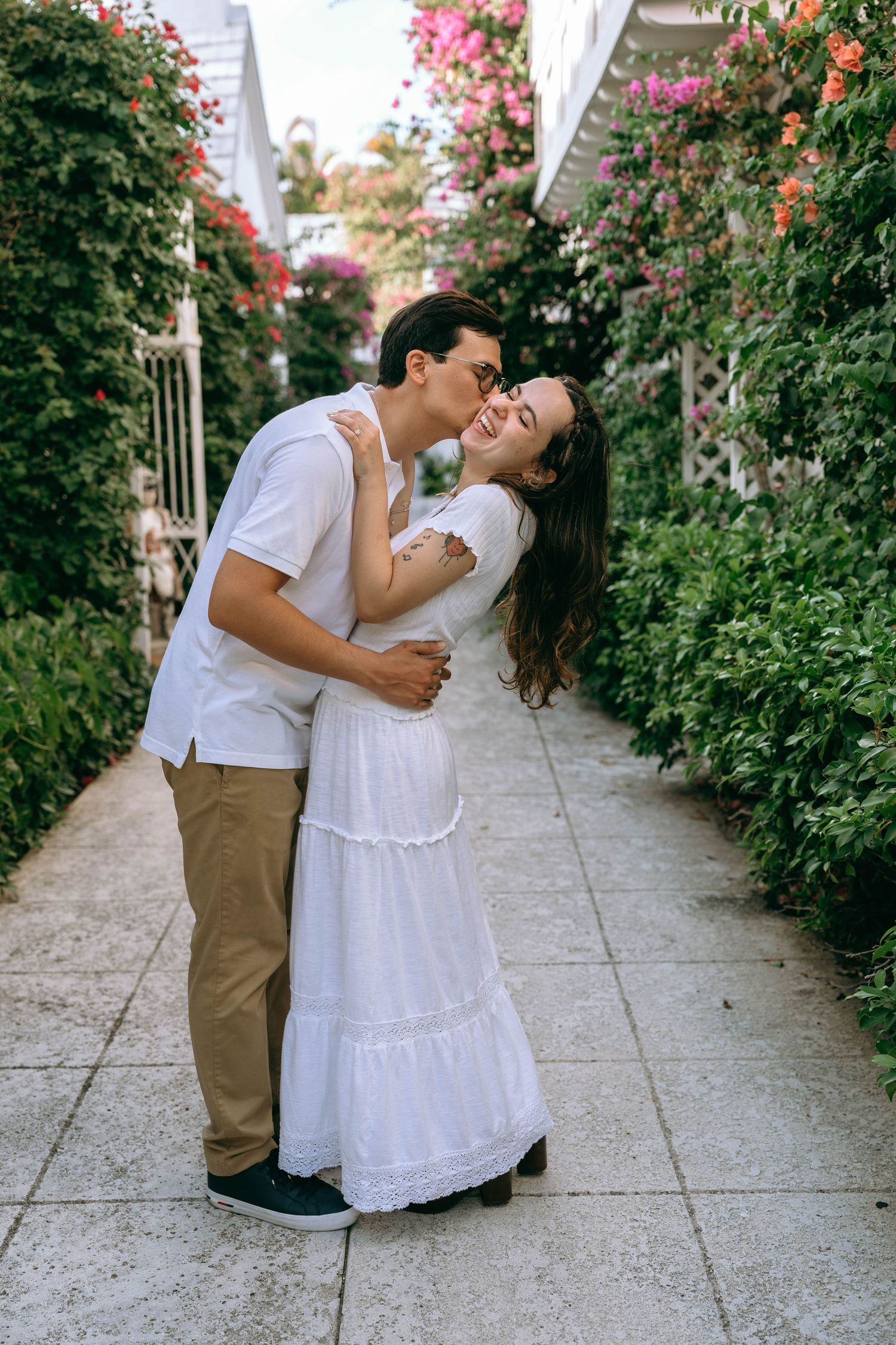 A couple embraces and shares a tender kiss in a lush garden pathway lined with colorful blooming flowers. Engagement photos on Worth Avenue