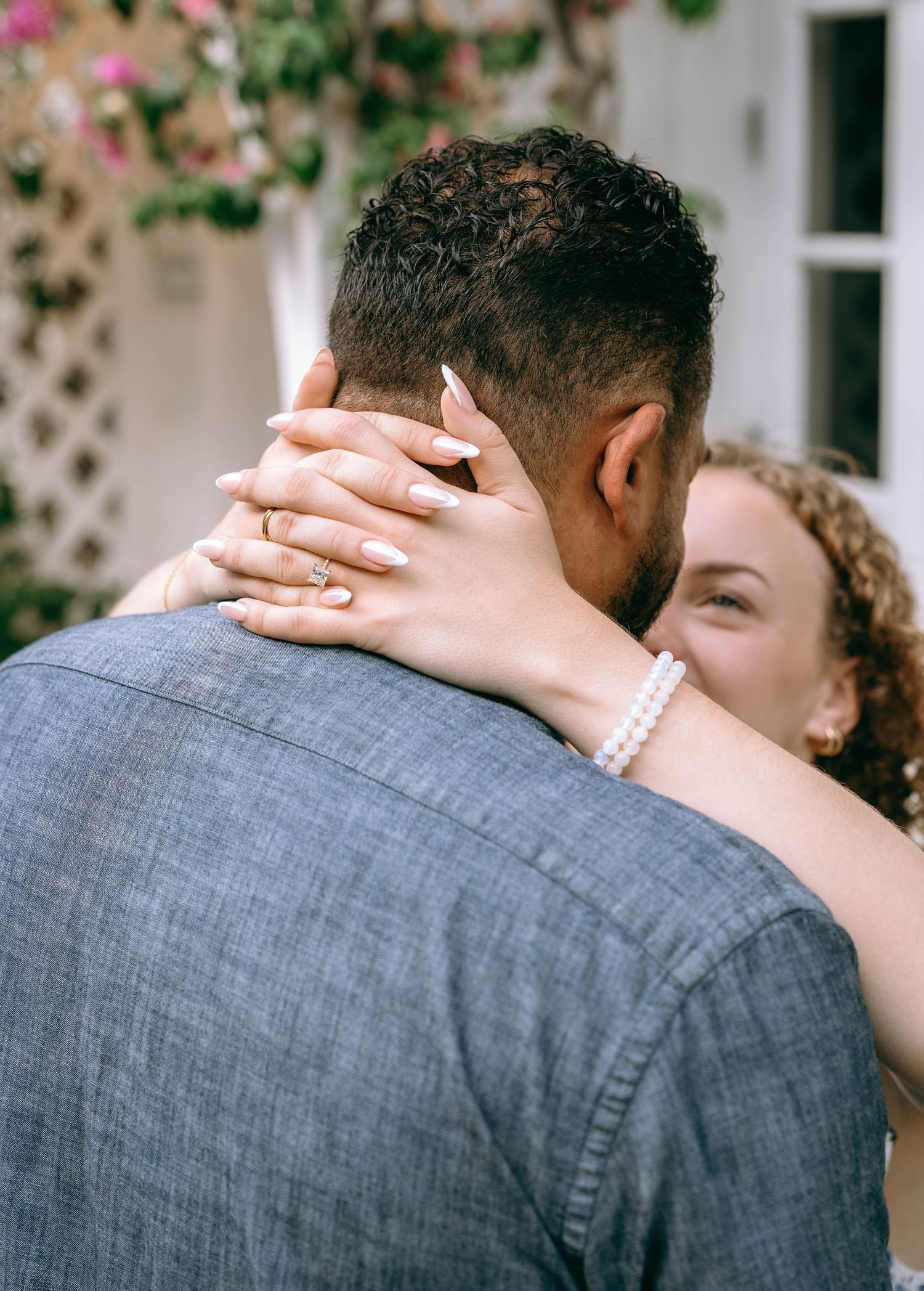 Close-up of woman with engagement ring and pearl bracelet embracing man in gray shirt outdoors. Engagement photos on Worth Avenue Palm Beach
