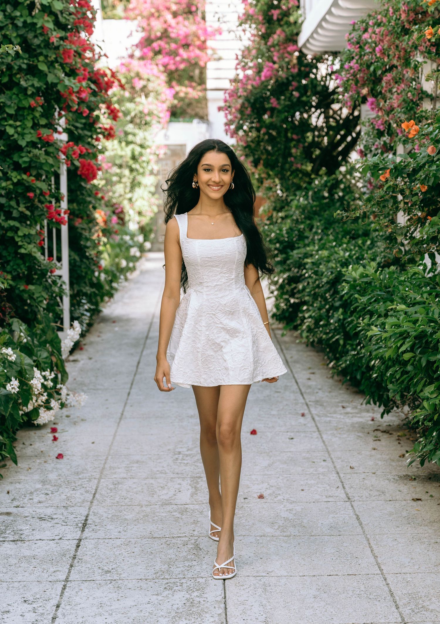 Young woman in white mini dress walking through a flower-lined garden pathway on Worth Avenue Palm beach.