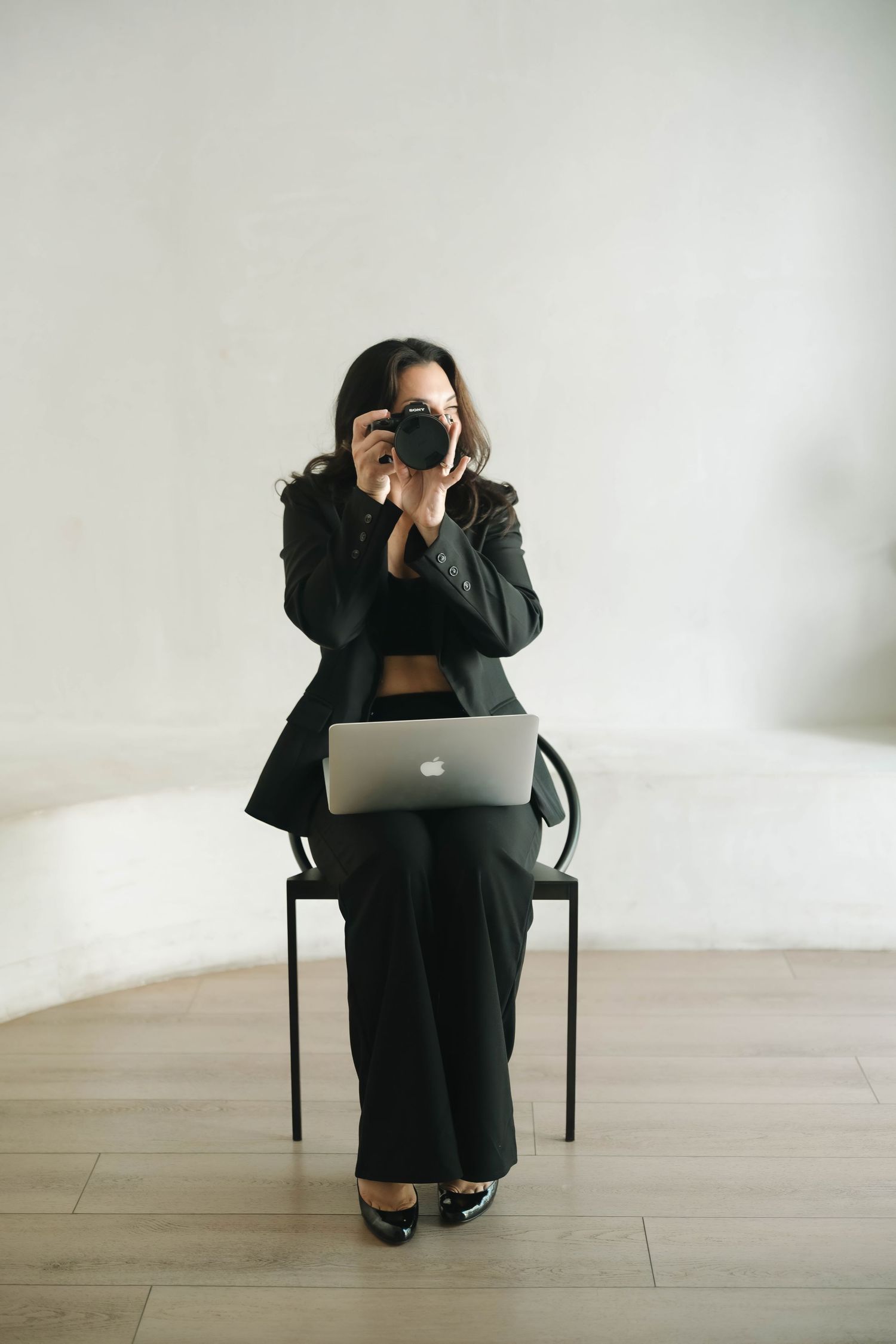 Person in black attire sitting on chair with laptop in minimalist white space.