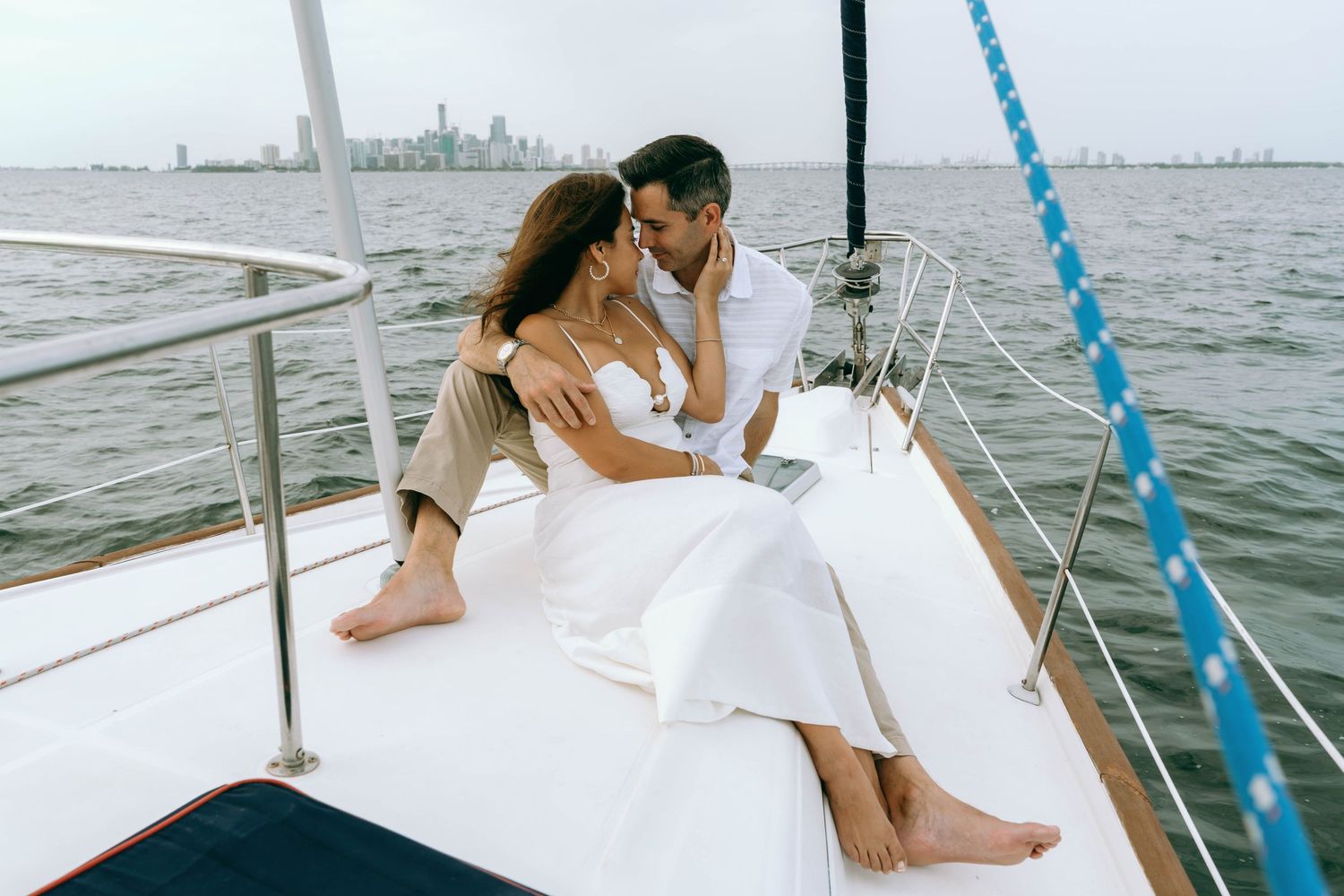 Romantic couple embracing on the bow of a white sailboat on calm waters with a city skyline in the background.