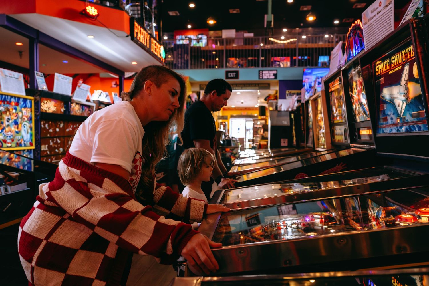 Adults and children playing pinball machines in a vibrant retro arcade game room with colorful cabinets.