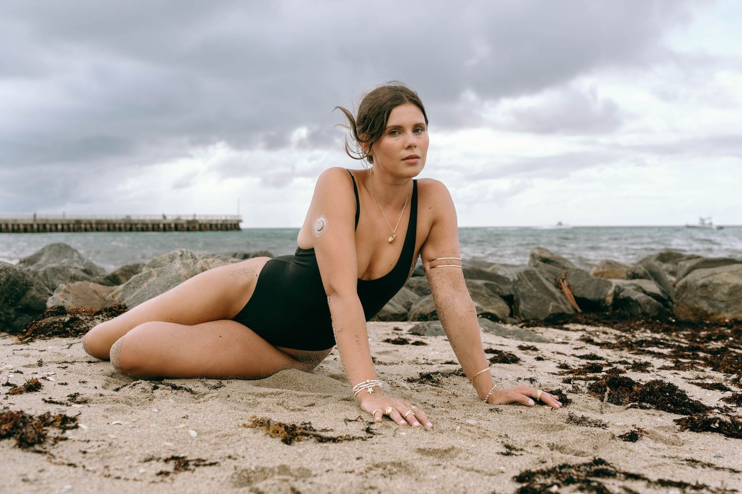 Woman in black swimsuit posing on sandy beach with rocky shoreline and cloudy sky backdrop.