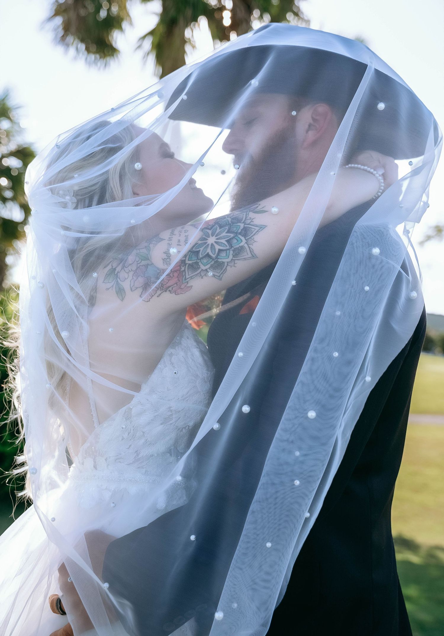 Wedding couple sharing intimate moment under flowing white veil outdoors.