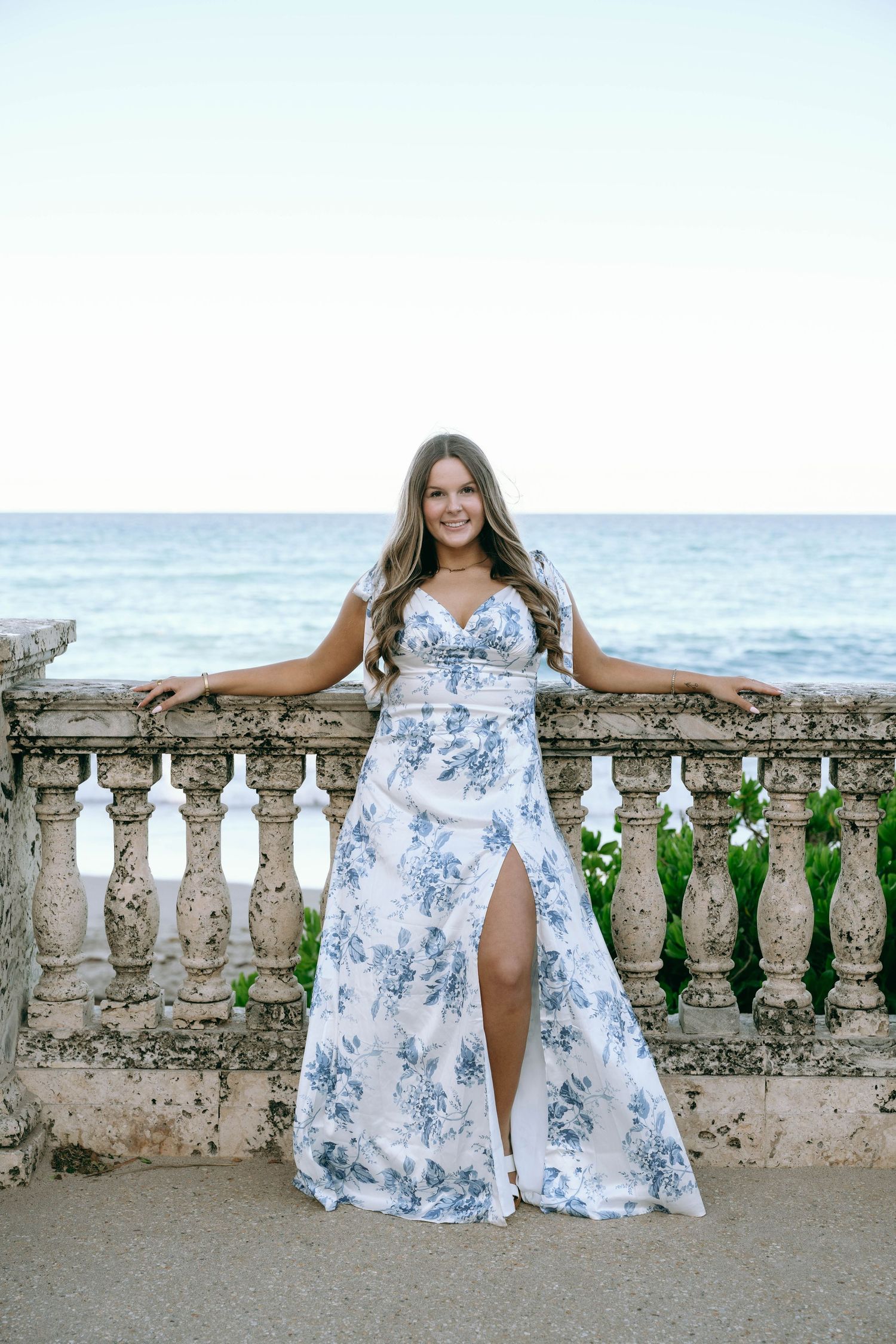 Woman in white and blue floral maxi dress with slit, posing at stone balustrade overlooking the ocean.