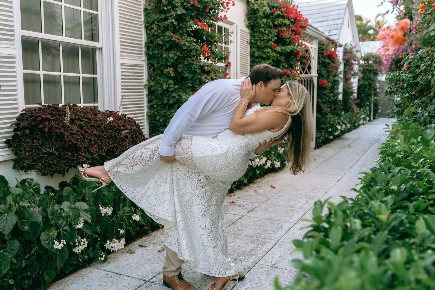 Couple sharing a romantic dip kiss along a flower-lined garden pathway.