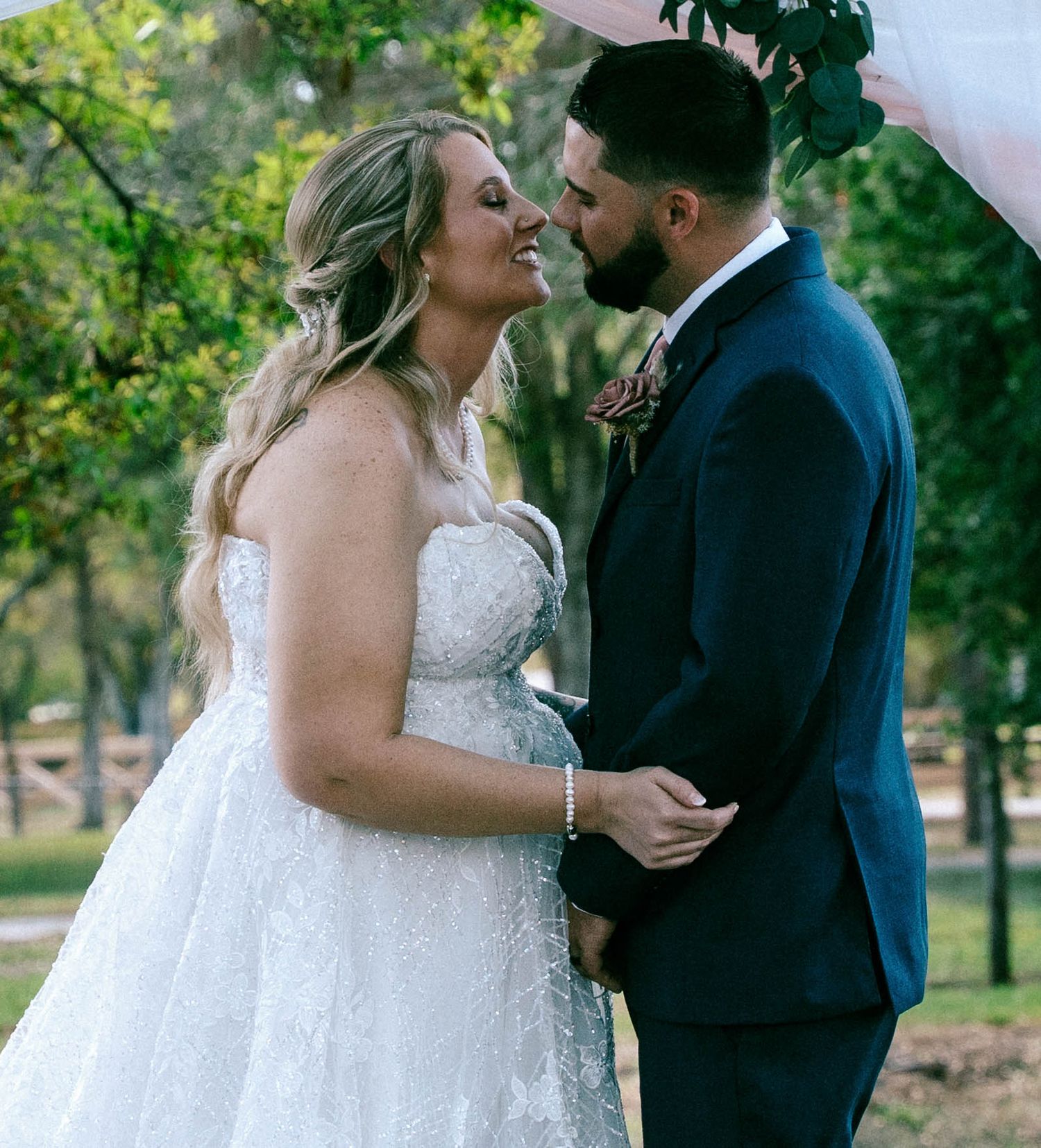 Bride and groom gazing lovingly at each other outdoors, bride in strapless lace gown, groom in navy suit.