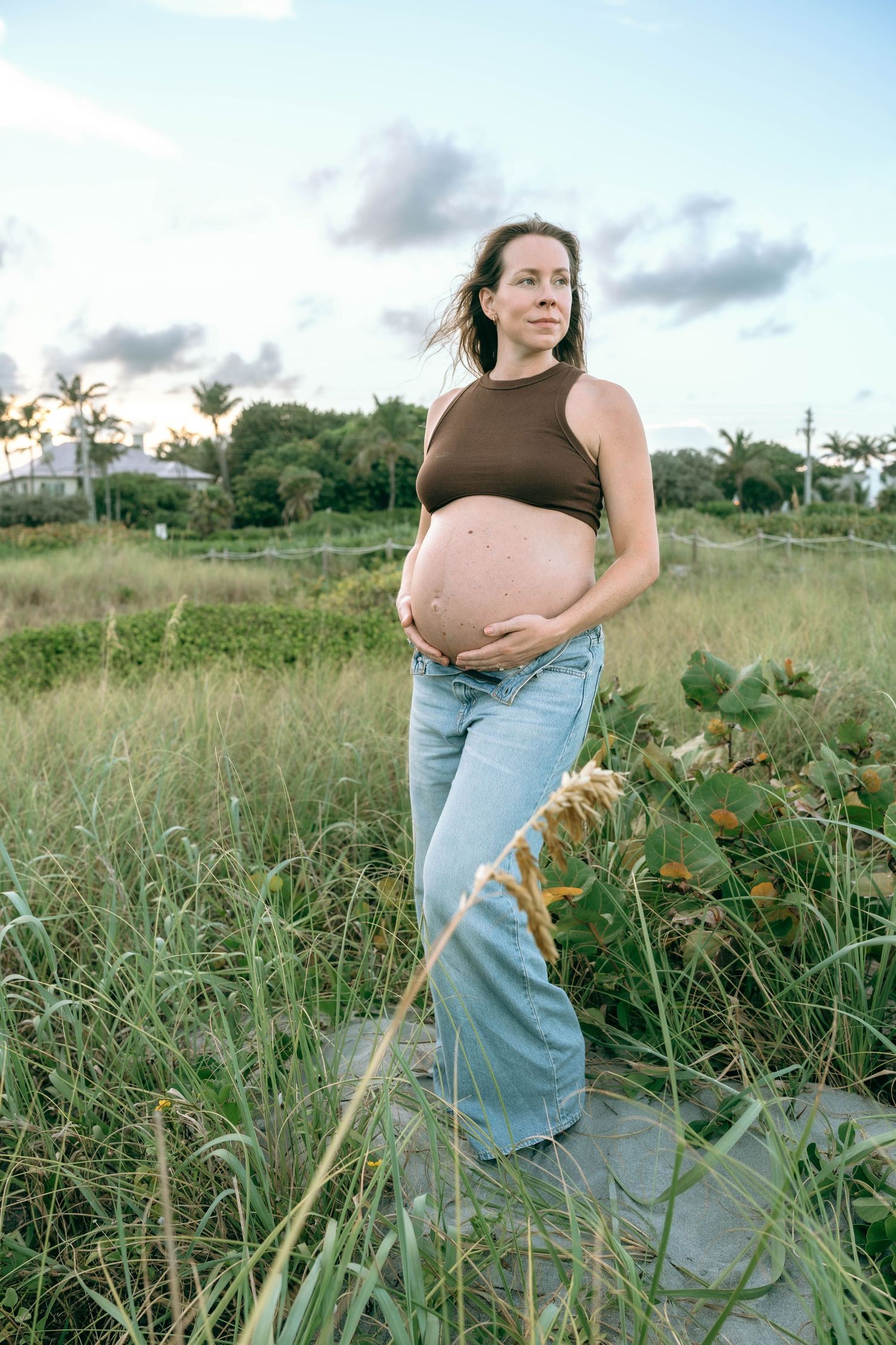 Pregnant woman in brown crop top and jeans cradling belly in lush green coastal field at dusk.