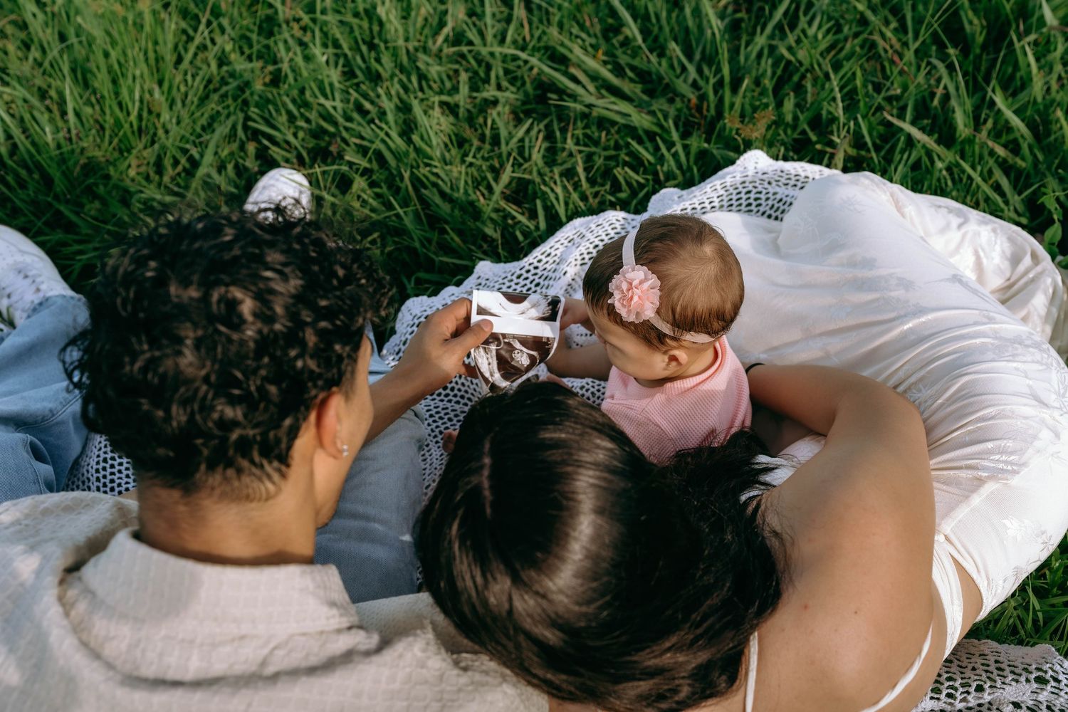 Two parents lying on grass with their baby in a pink outfit, sharing a tender family moment outdoors.