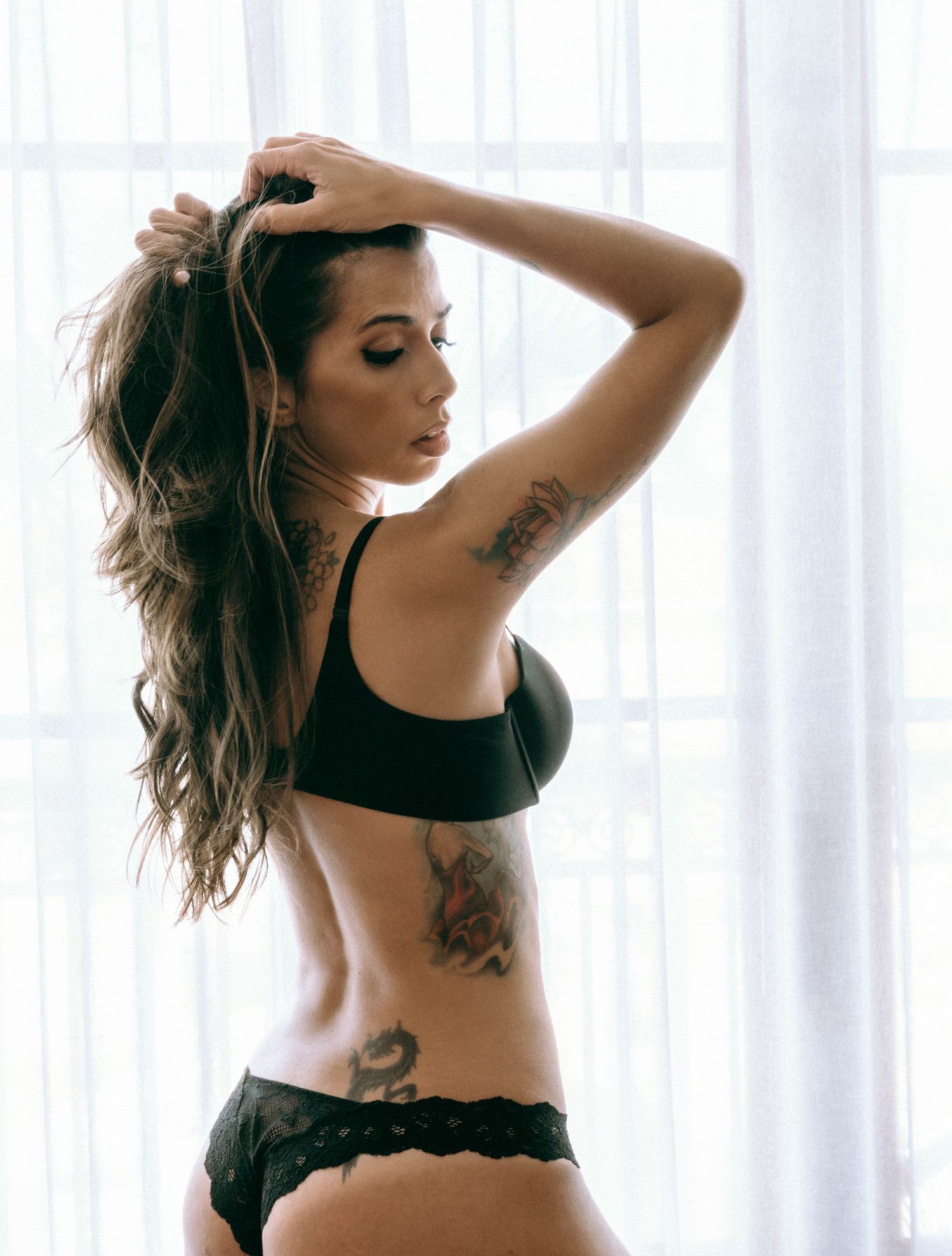 Woman in black lingerie with tattoos posing by sheer curtains.