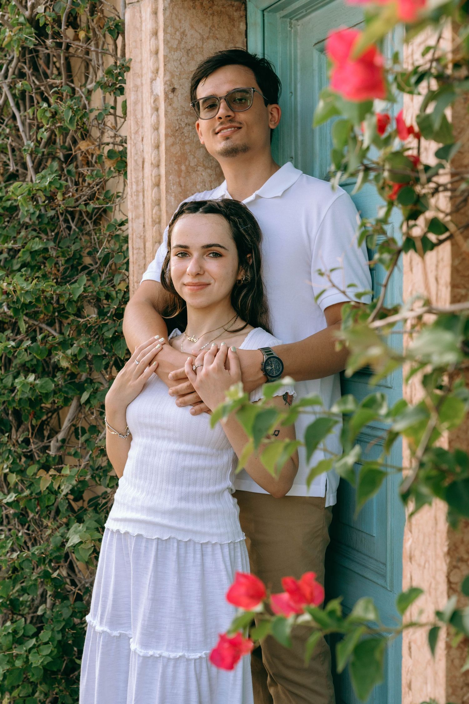 A couple poses together near a teal door surrounded by lush greenery and red flowers, woman in white dress. Engagement photos on Worth Ave