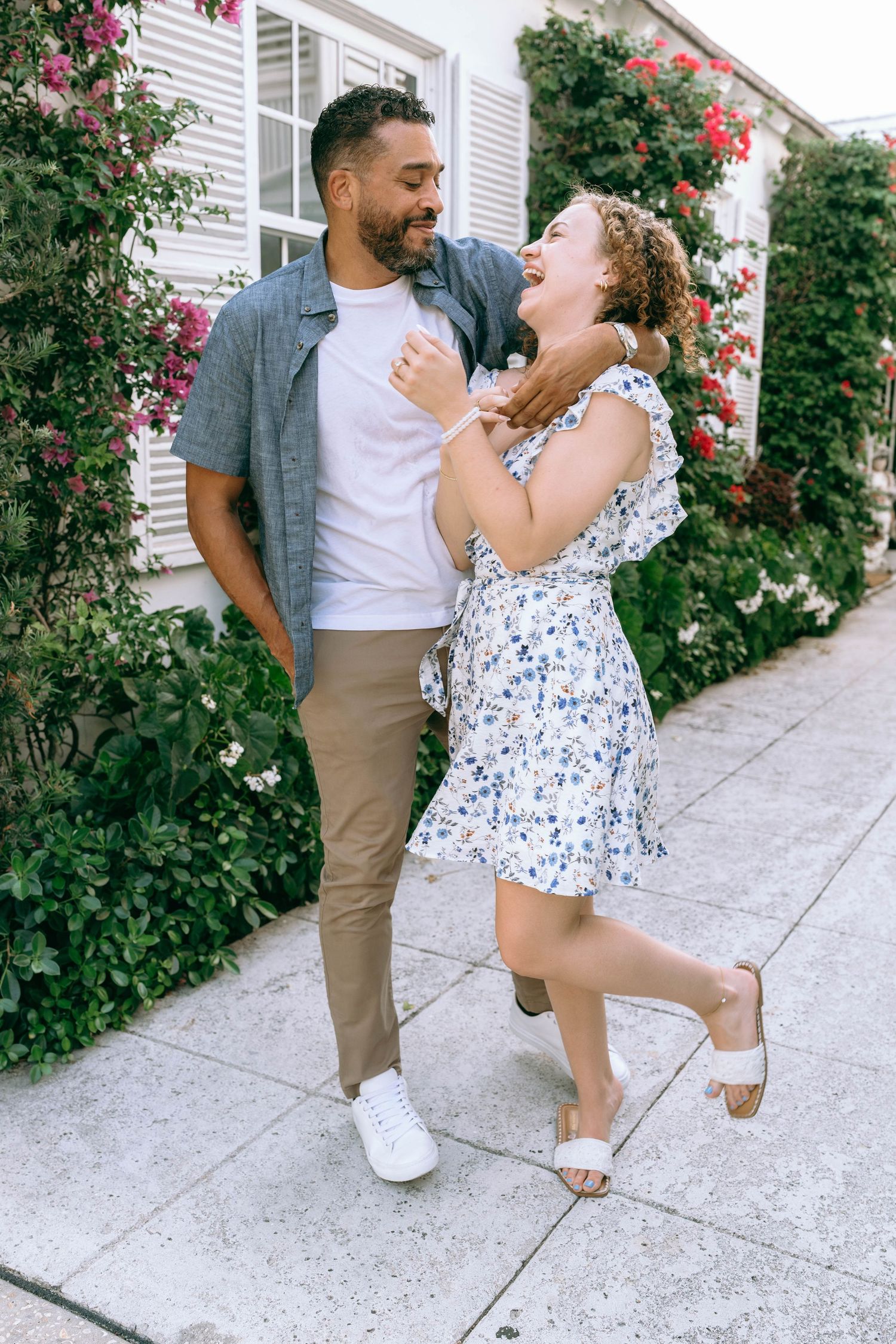 Happy couple laughing and dancing together on a sidewalk surrounded by blooming rose bushes. Engagement photos on Worth Avenue in Palm Beach