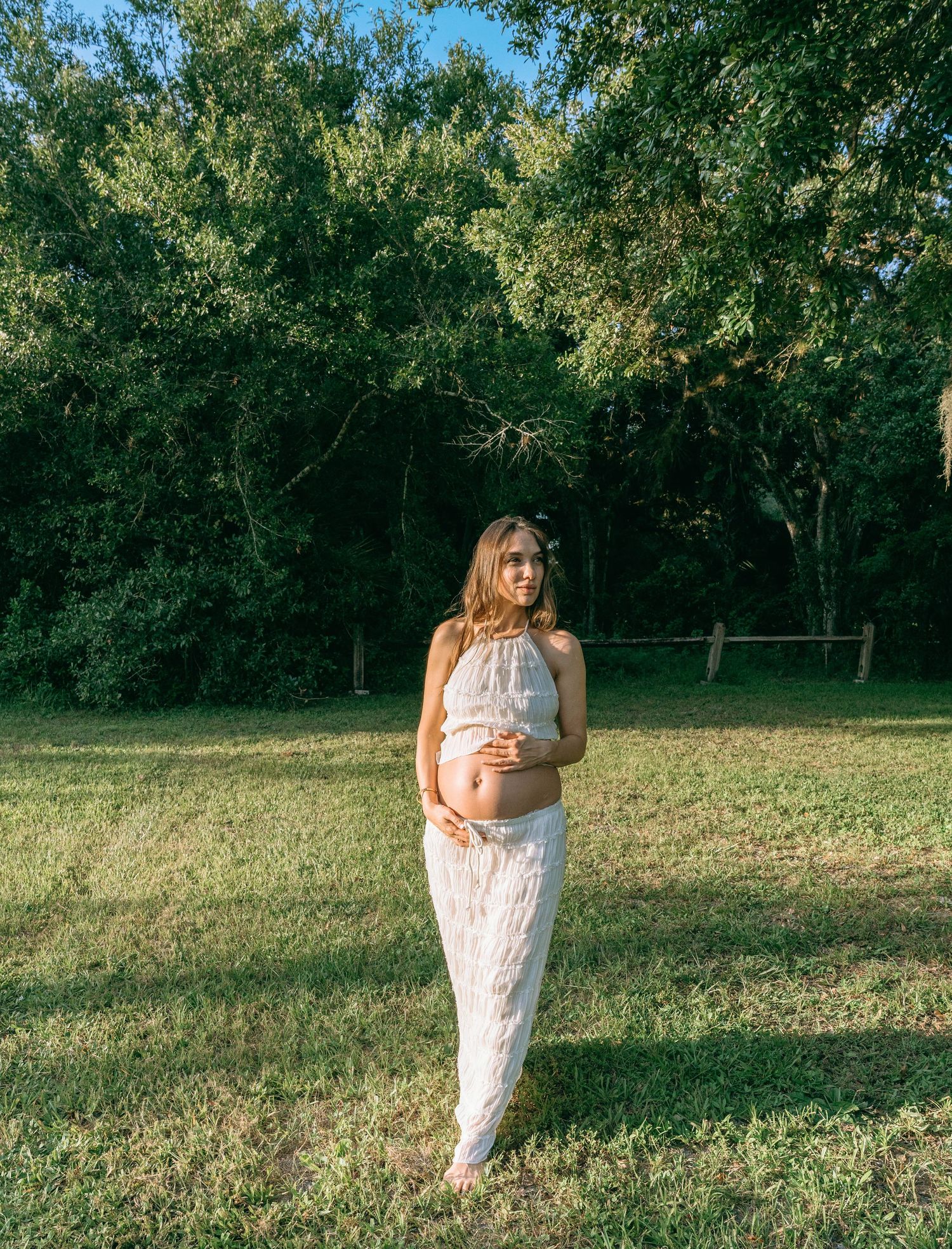 Maternity photoshoot in a sunlit field with a flowing white dress and natural greenery backdrop.