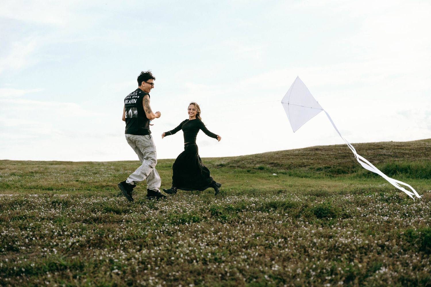 Two people run through a grassy field flying a kite on a cloudy day.