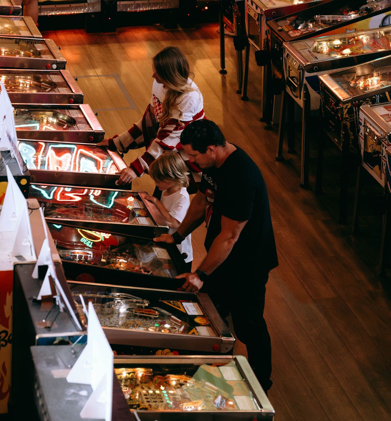 People playing pinball machines in a retro arcade hall with wooden floors and rows of colorful illuminated games.