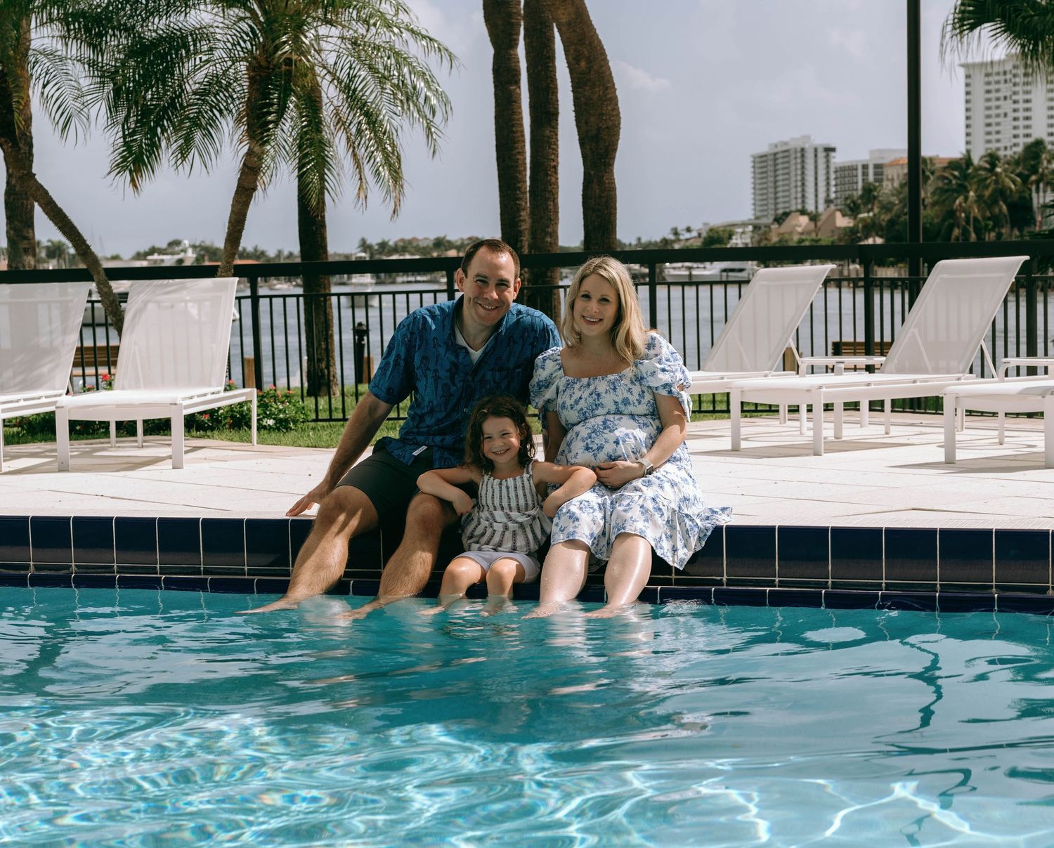 Family of three sitting poolside with feet in water, palm trees and city skyline in background on cloudy day.