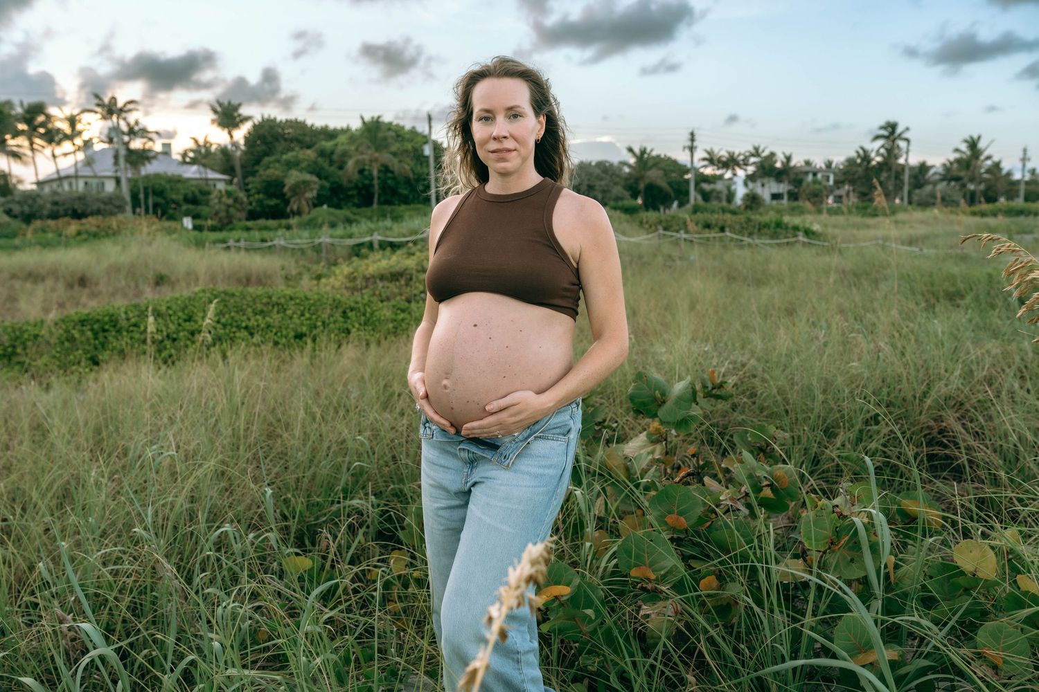 Pregnant woman in brown crop top and jeans standing in green field at dusk.