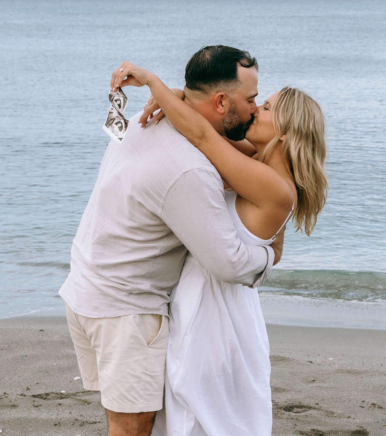 Couple kissing on a sandy beach, woman in white dress holding shoes, man in white shirt and khaki shorts, ocean background.