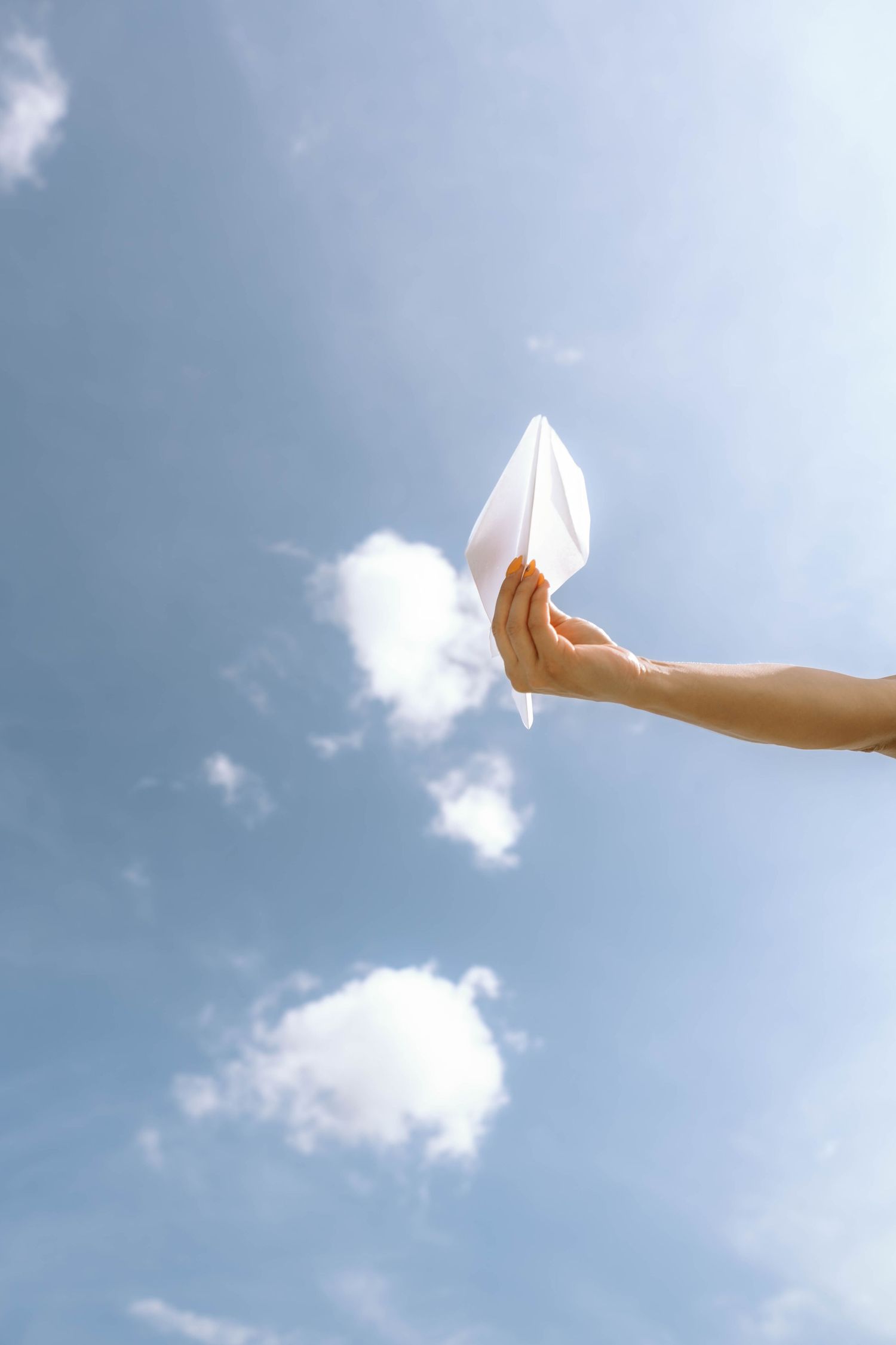 Hand holding a white paper airplane against a bright blue sky with fluffy clouds on a sunny day.