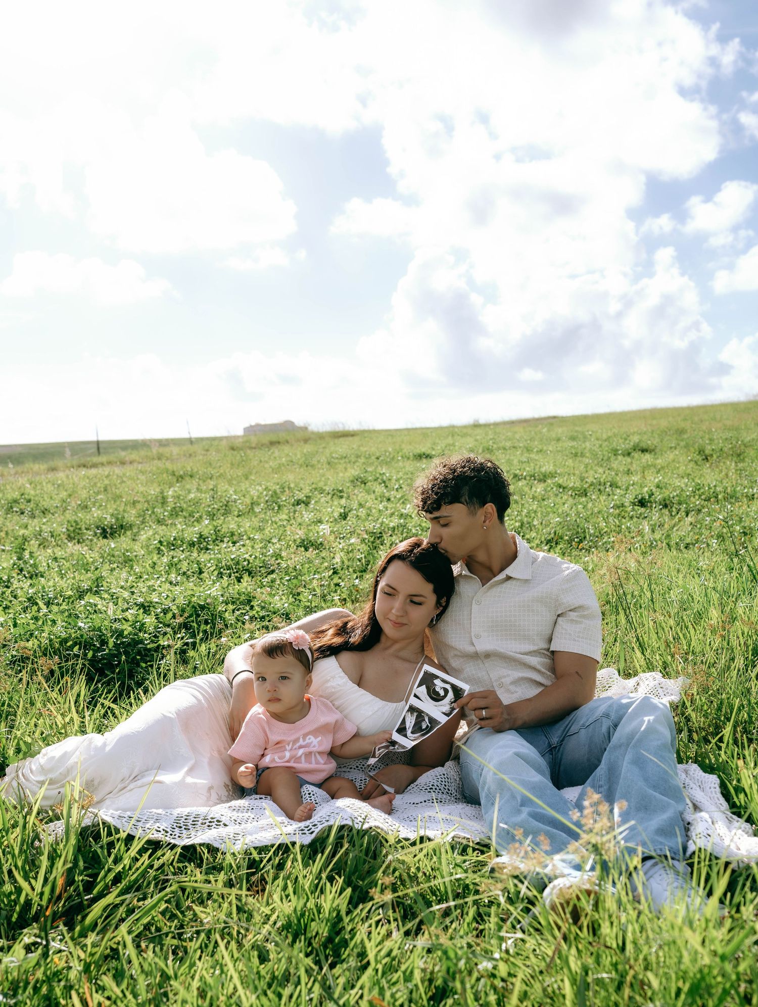 A young family sits on a blanket in a green field holding an ultrasound photo, announcing a pregnancy.