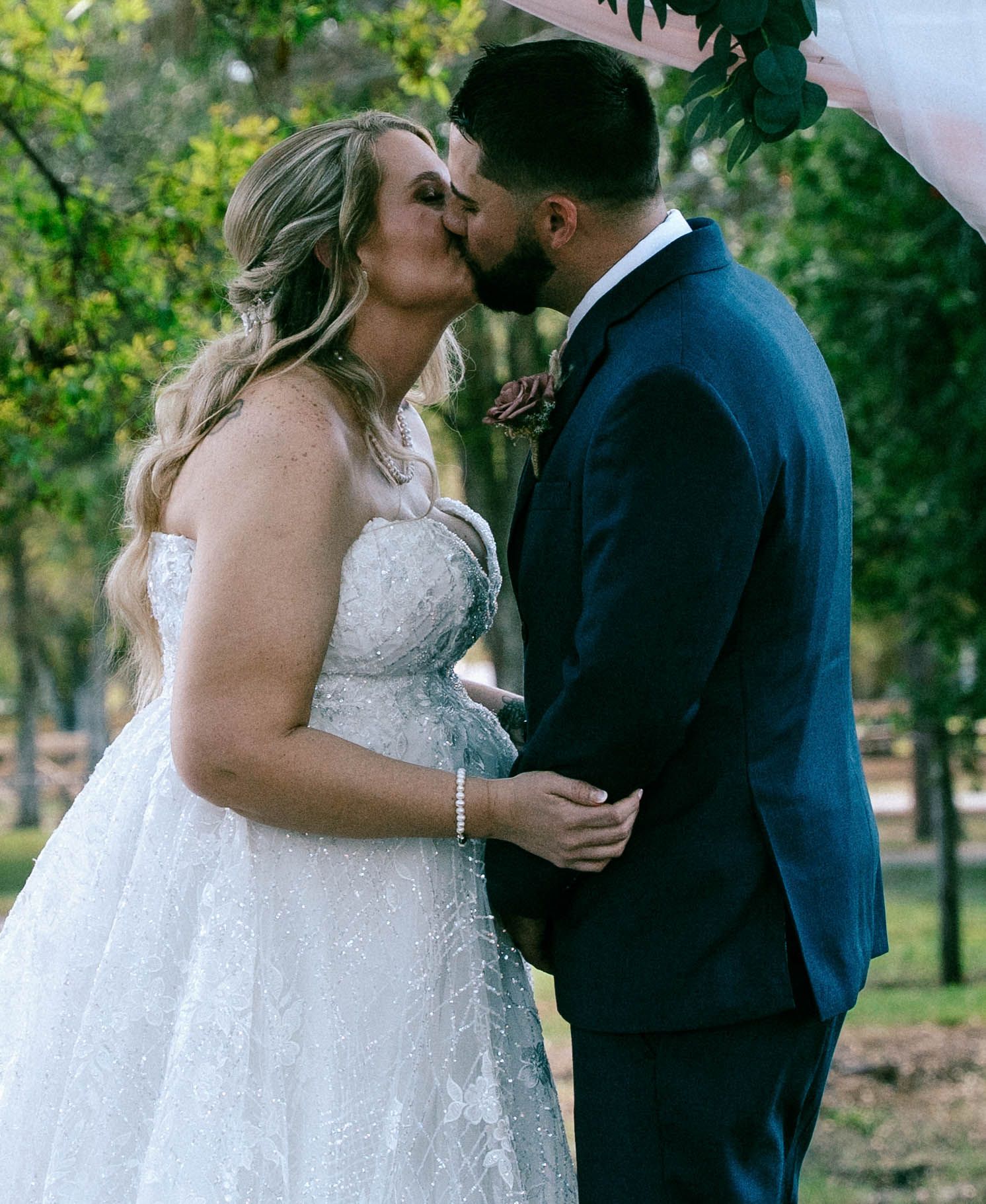 Bride and groom sharing first kiss at outdoor wedding ceremony, bride in lace ballgown, groom in navy suit.