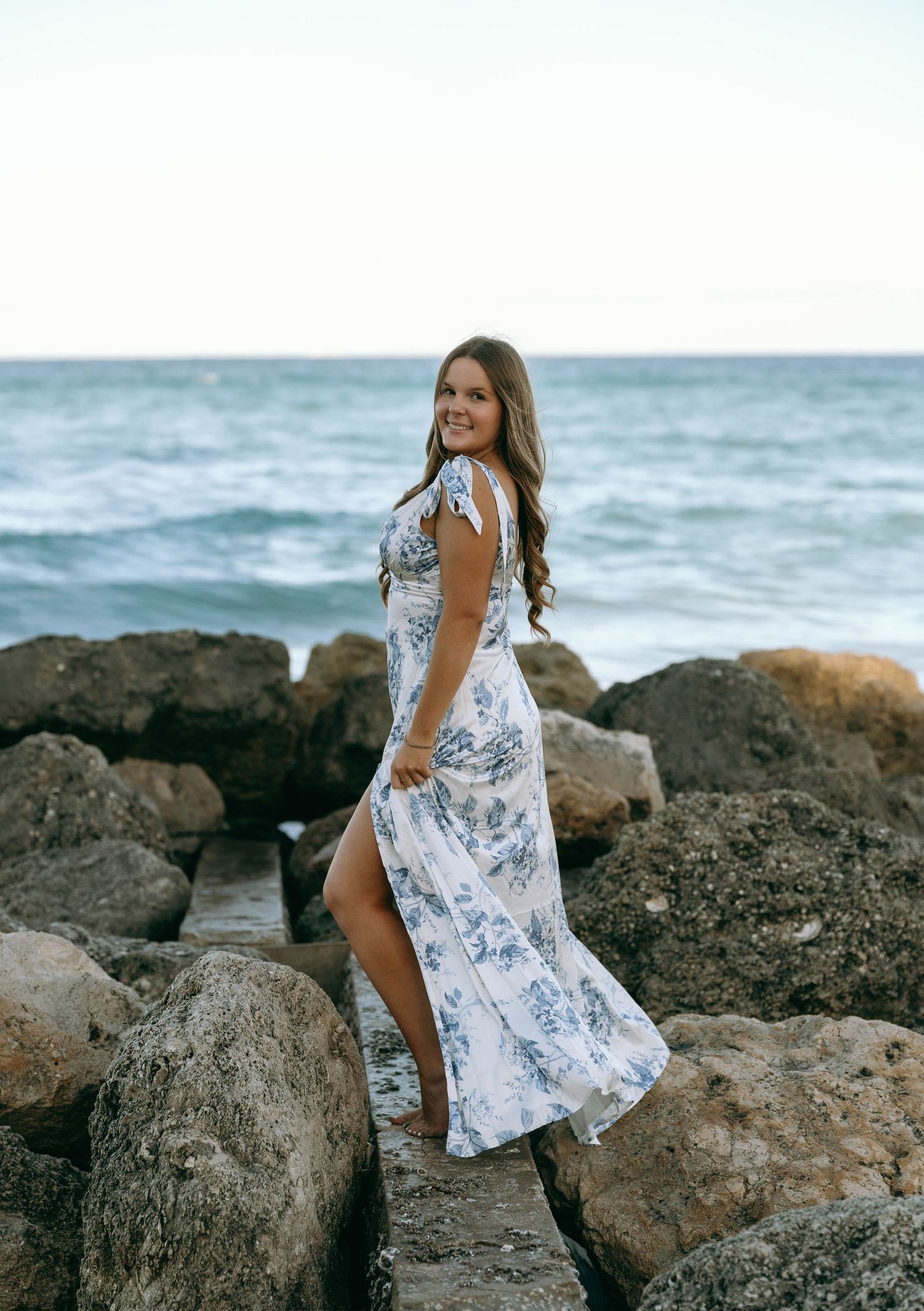 Woman in white blue floral maxi dress standing on rocky oceanside shoreline.