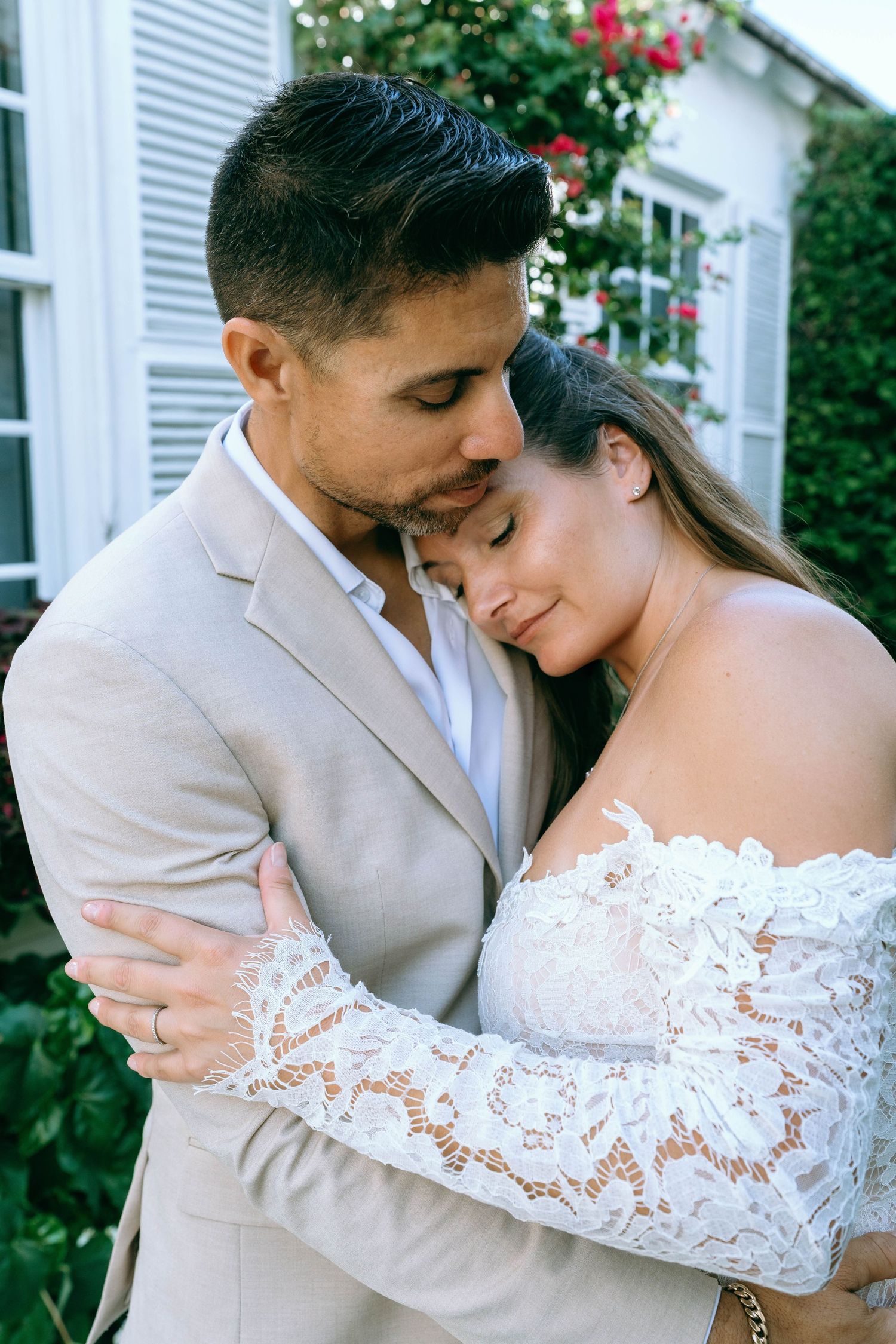 Newlyweds share a tender embrace, bride in off-shoulder lace dress and groom in beige suit near floral backdrop.