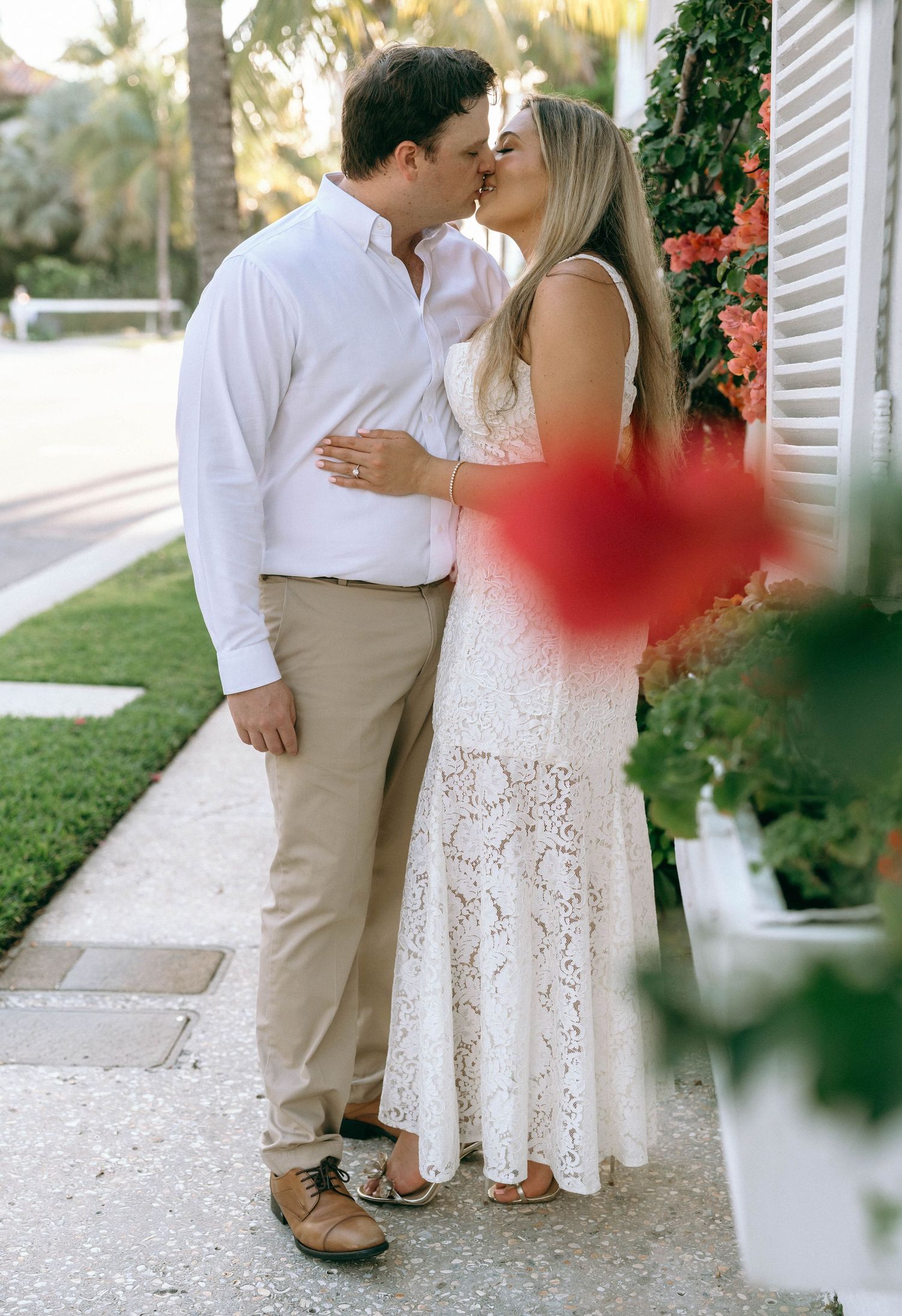 Couple sharing a kiss outdoors in elegant white lace dress and khaki pants.