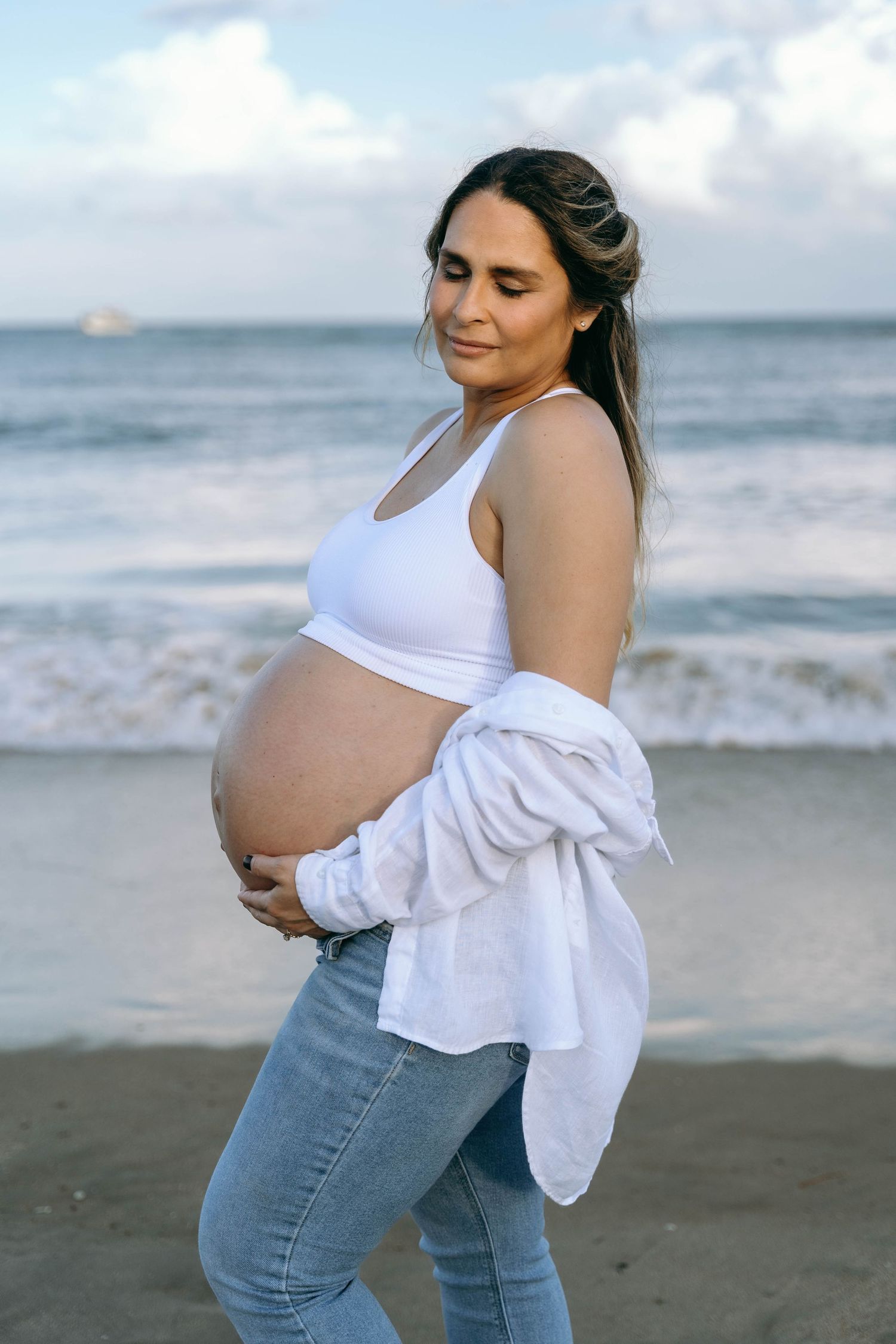 Maternity photo at the beach showing a pregnant woman in jeans and white top against ocean waves.