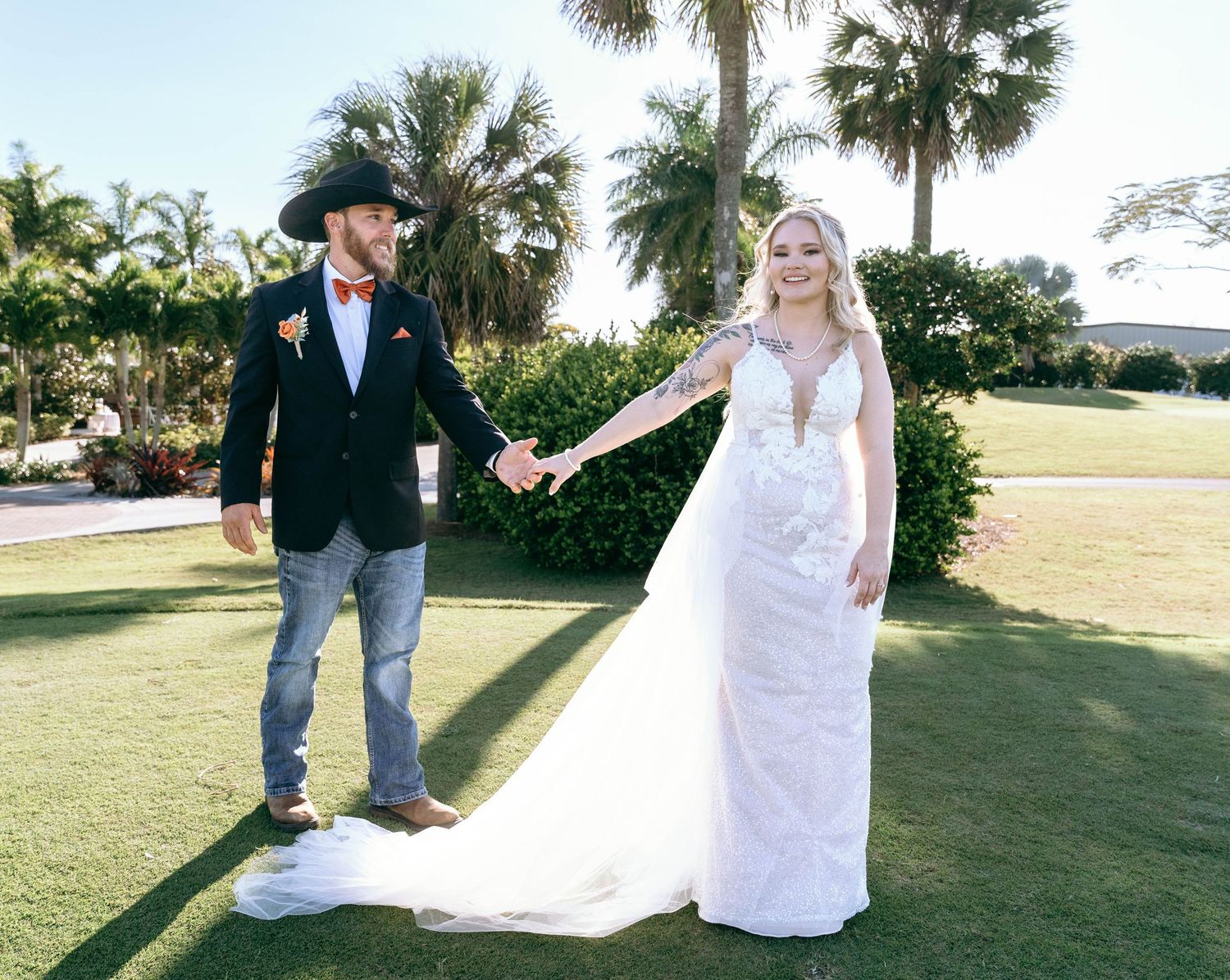 A couple poses on a sunny golf course with palm trees, the bride wearing a flowing white lace gown.
