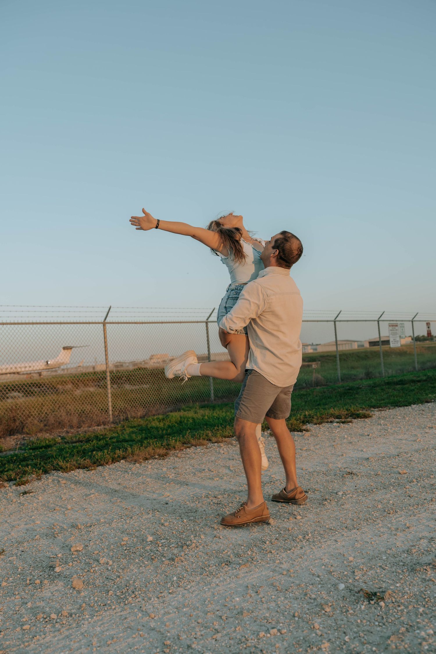 A man lifts a joyful woman with outstretched arms on a gravel path near a fence at sunset.