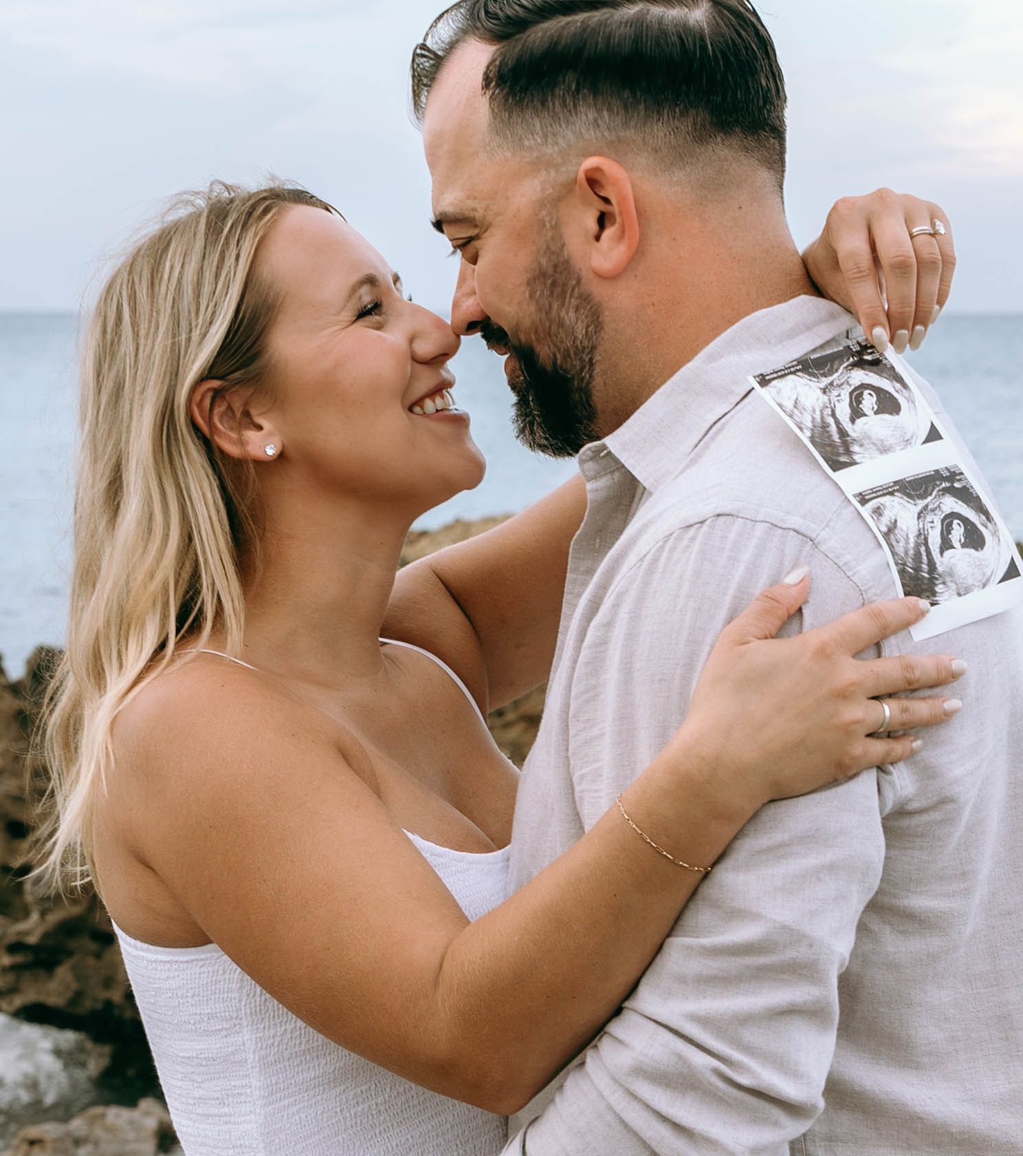 Happy couple embracing outdoors holding ultrasound photos, announcing pregnancy near rocky ocean shore at sunset.