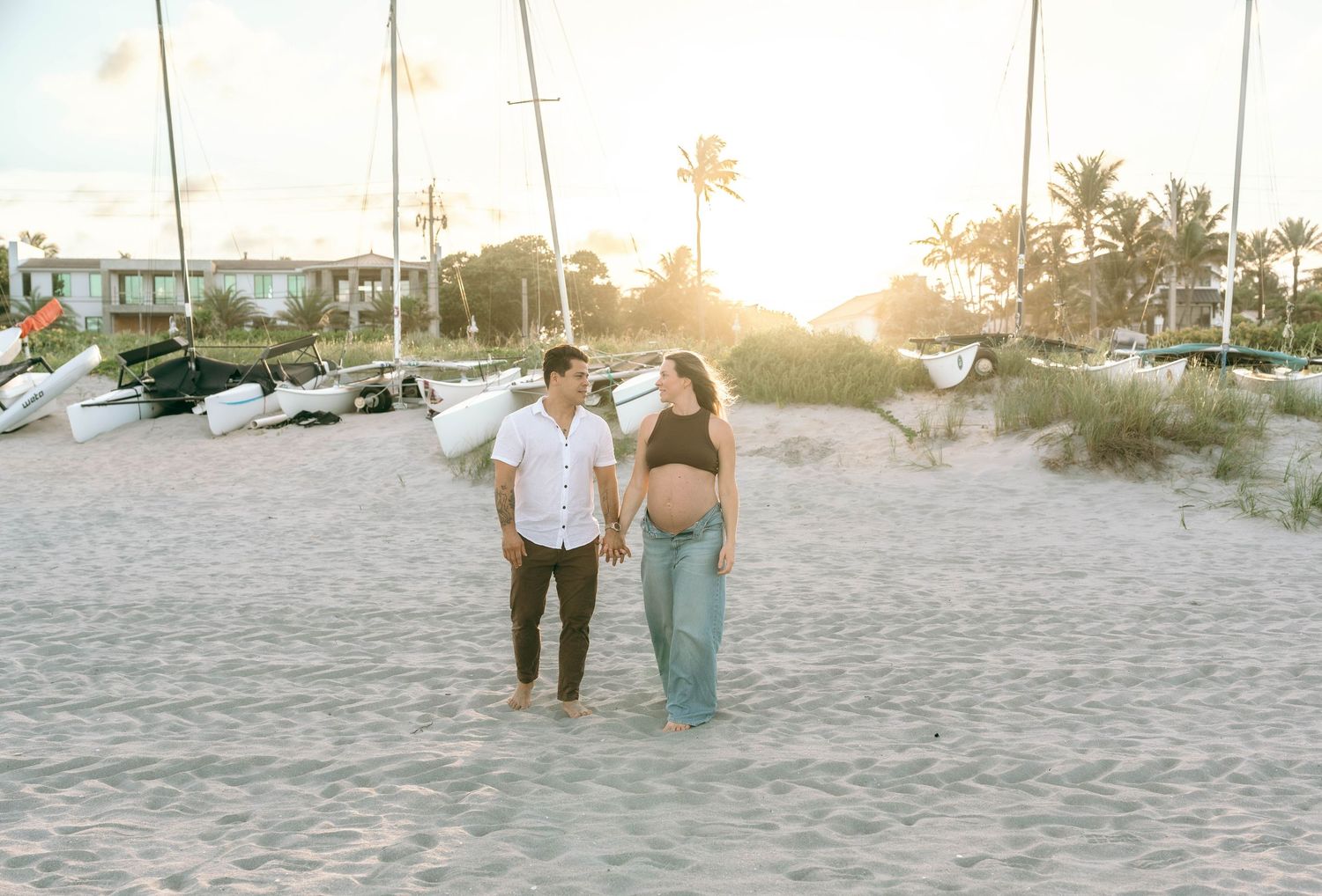 Pregnant couple walking on sandy beach at golden sunset with palm trees and boats in background.