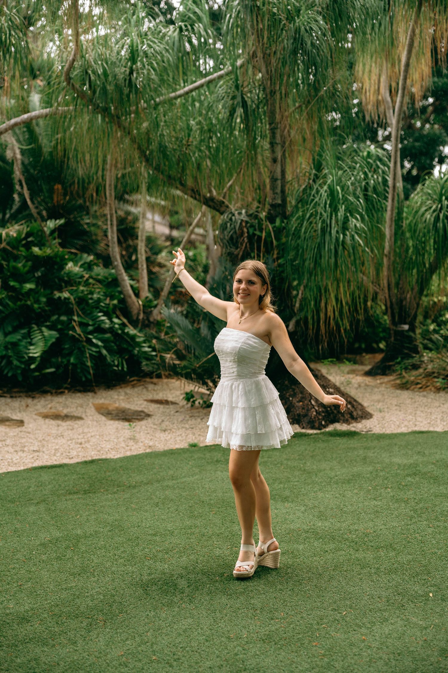 Young woman in white tiered strapless dress dancing near tropical palm trees on green grass.
