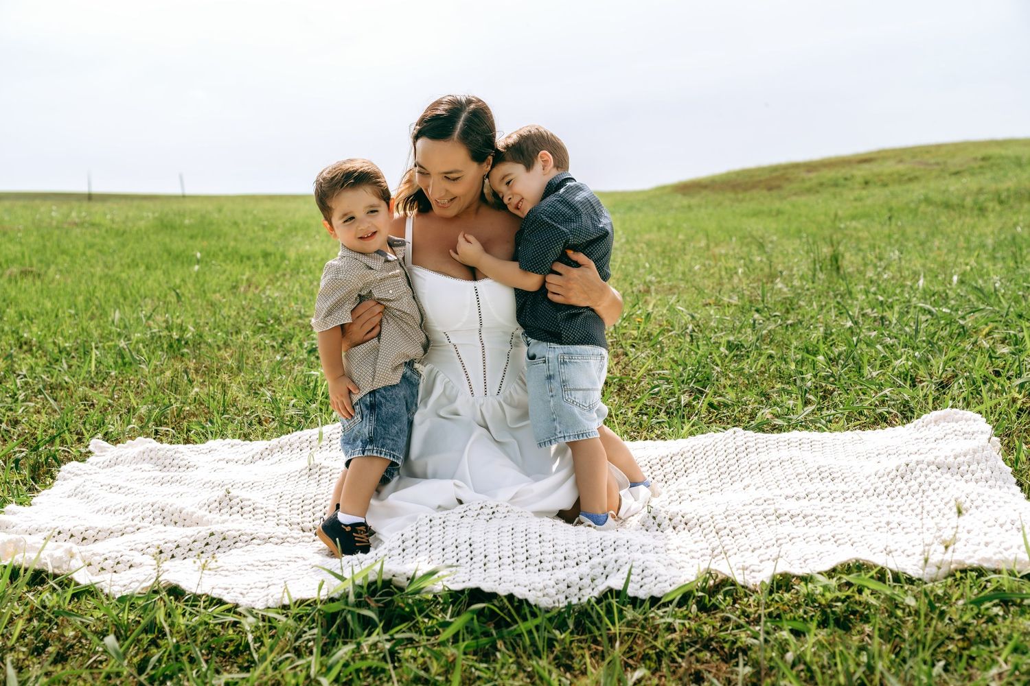 A mother sits on a white blanket in a green field, hugging her two young sons in a heartwarming outdoor portrait.