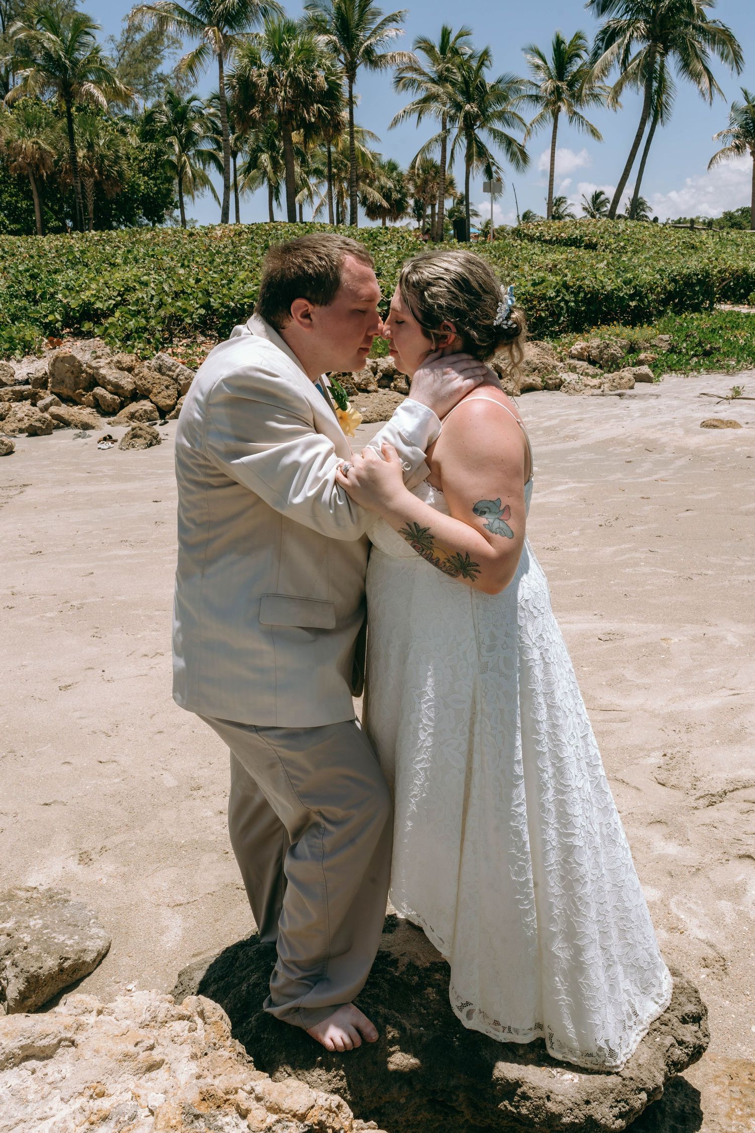 Bride and groom sharing a romantic kiss on a tropical beach, dressed in white lace gown and tan suit.