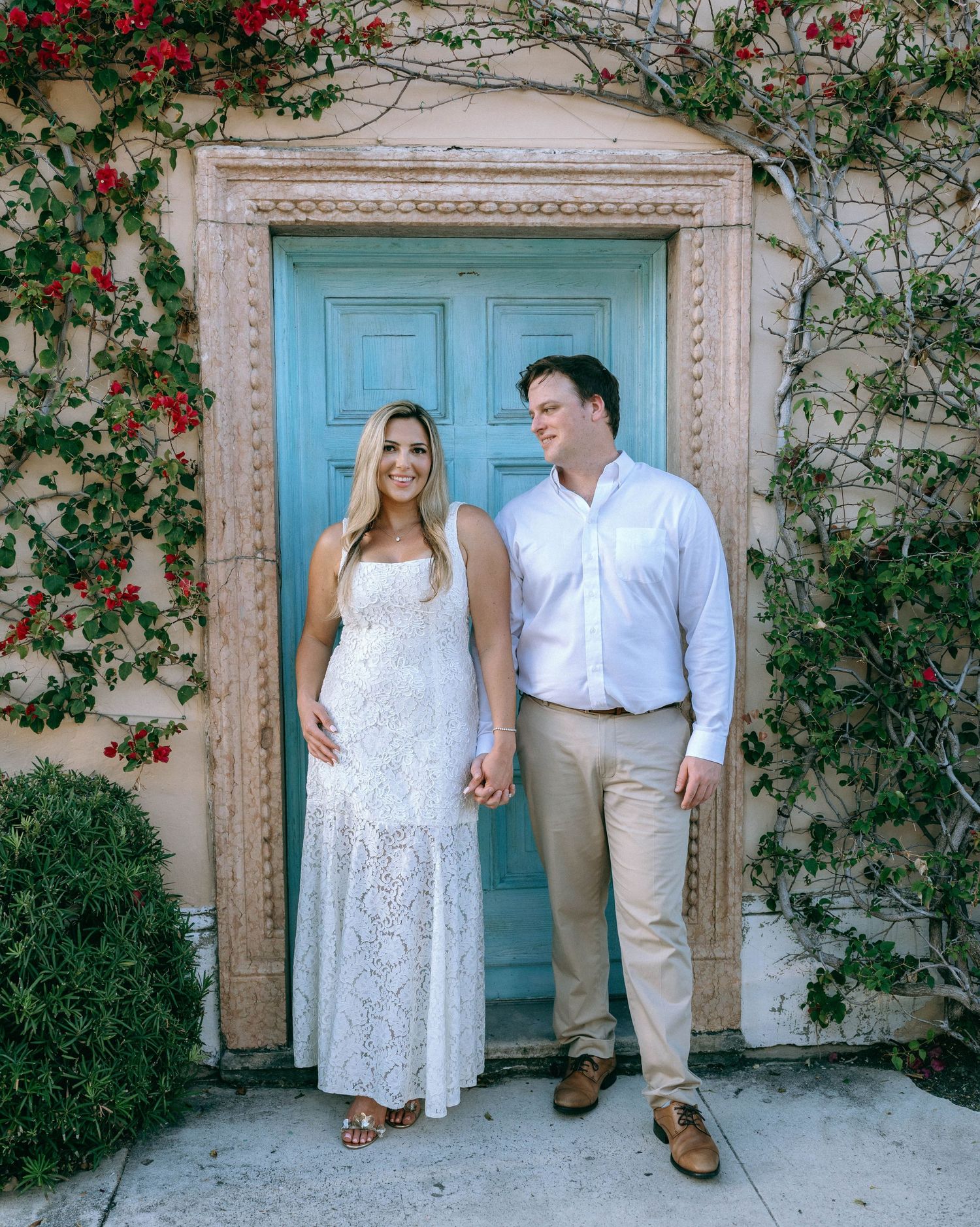 Smiling couple holding hands in white outfits before a turquoise door framed by red flowering vines.